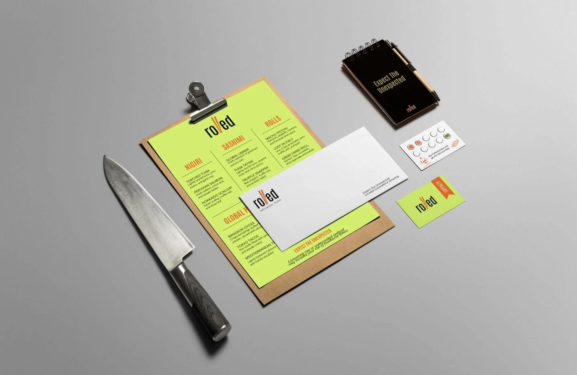 A menu on a clipboard with a knife beside it, a small notepad, business cards, and a card with orange and gray designs on a gray surface.