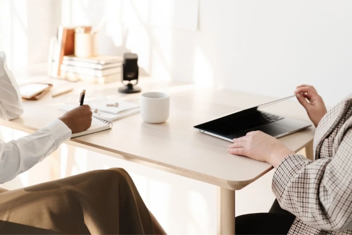 Two people sitting at a light-colored desk at a marketing agency during a meeting; one person is taking notes with a pen, and the other is using a stylus on a laptop.
