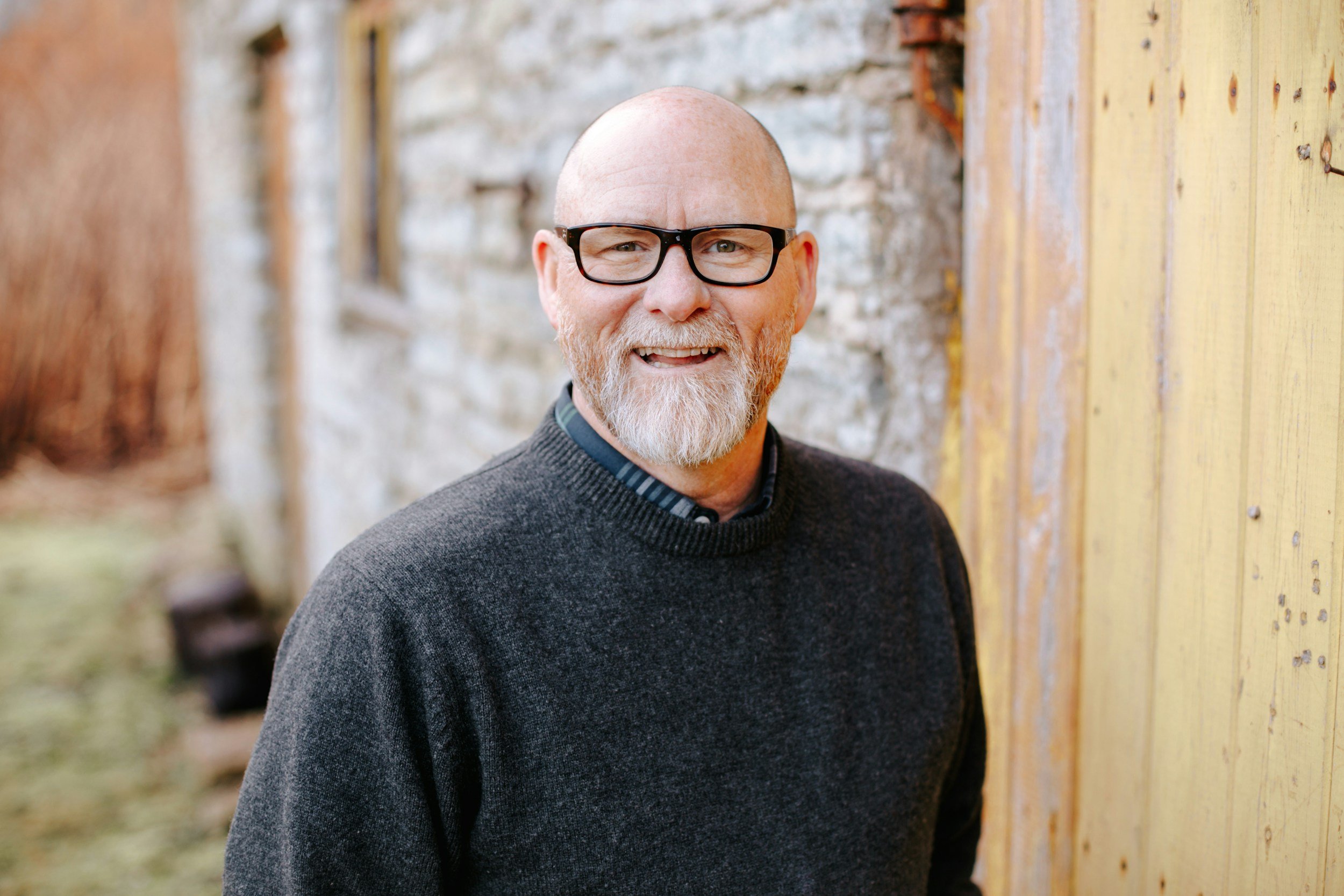 A smiling bald man with glasses and a beard, wearing a dark sweater, standing outdoors near a rustic yellow wooden building and a gray brick wall.