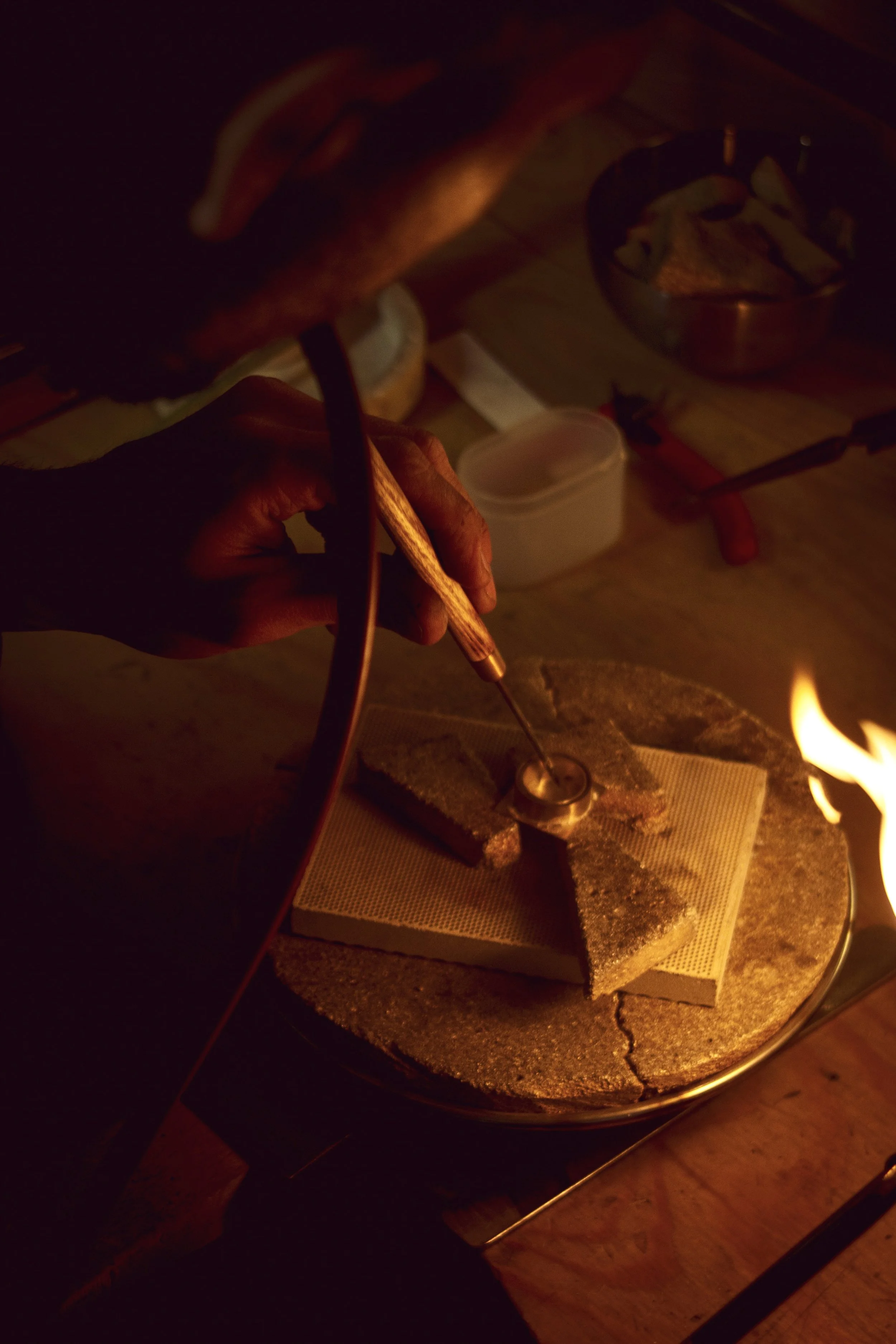A person working on a jewelry piece using a small torch and tools on a workbench, illuminated by a small flame.