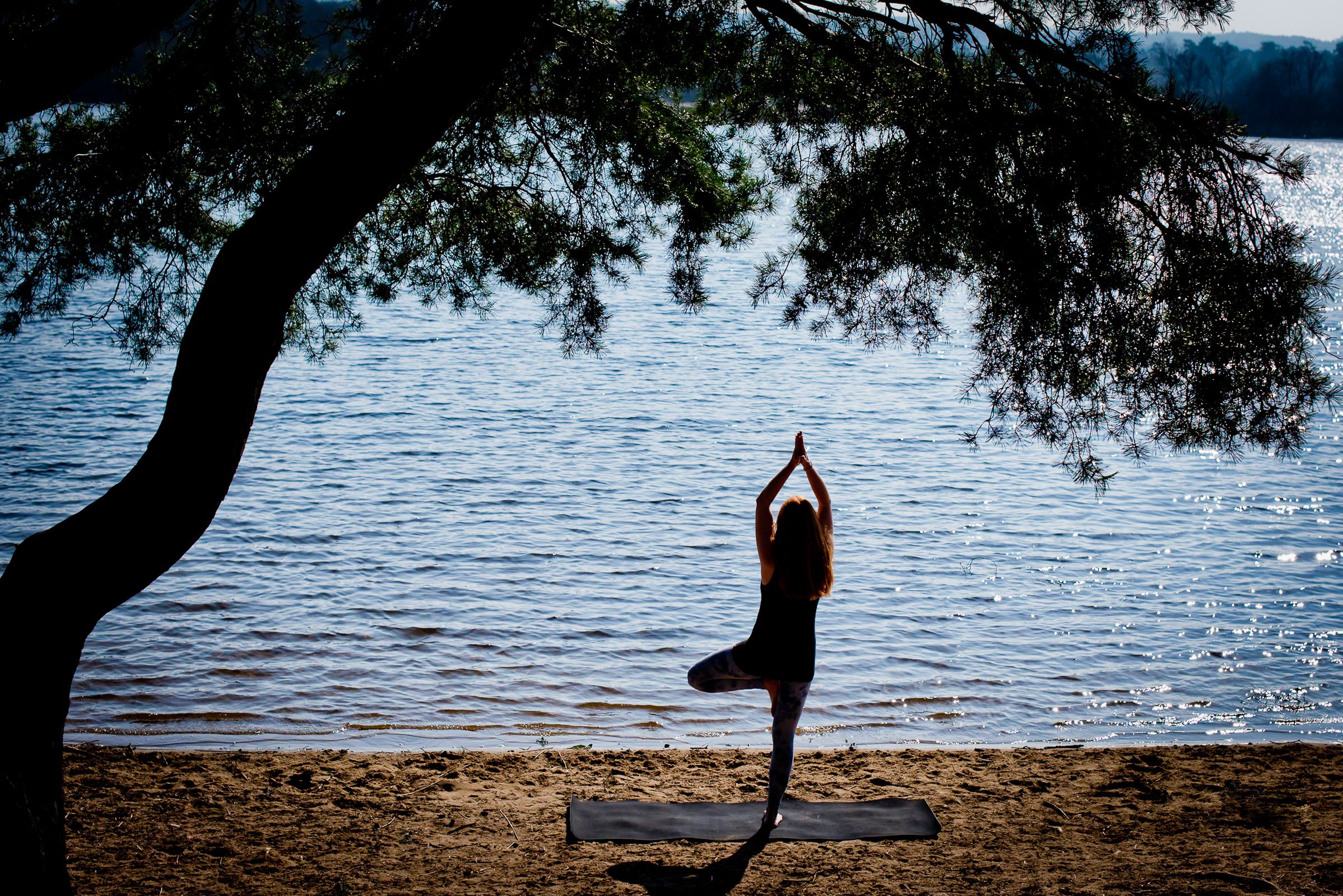 Sophie practicing yoga on a mat at the beach, performing the tree pose, with a large tree overhead and calm water in the background.