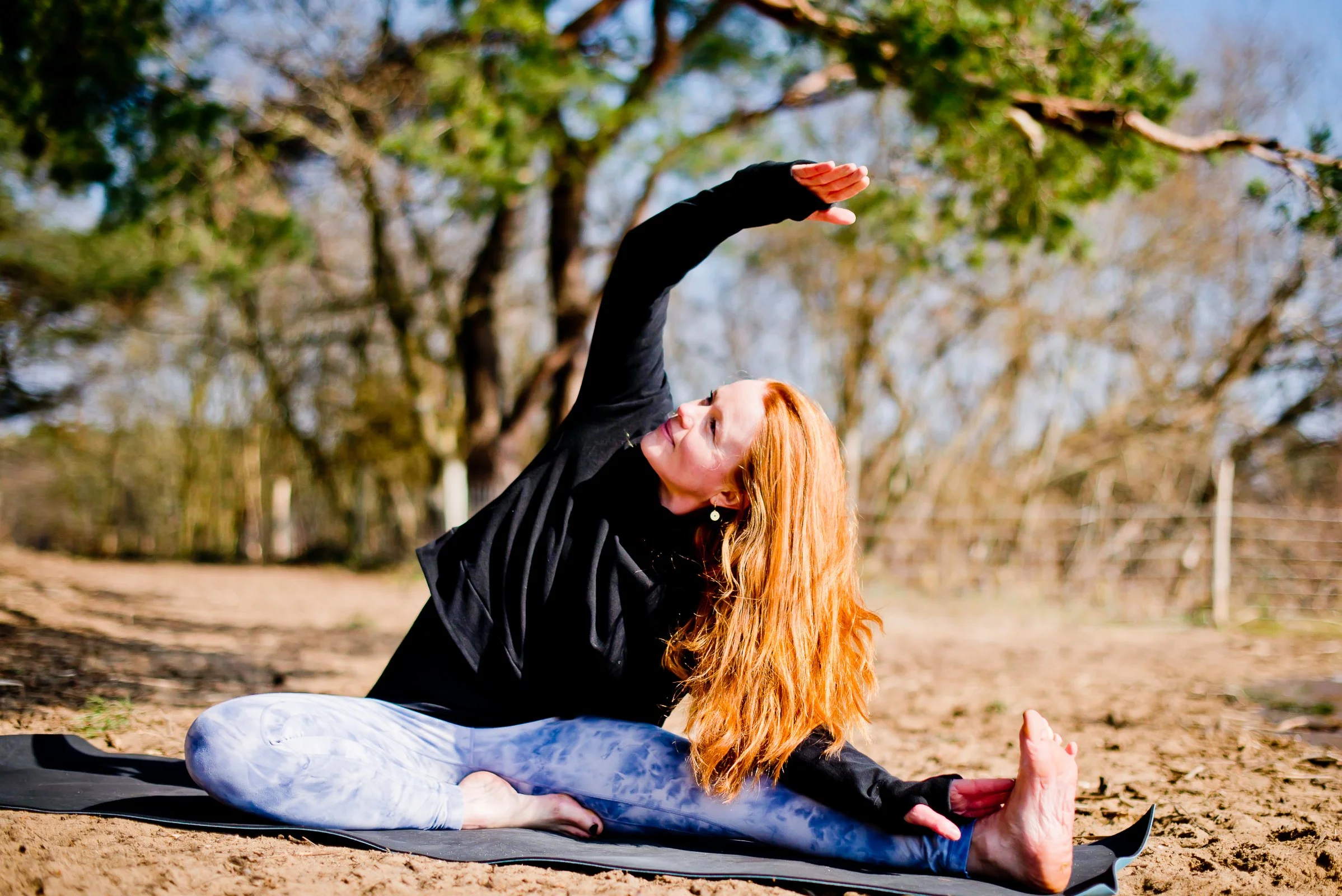 Sophie doing a seated side stretch yoga pose outdoors on a sunny day with trees in the background.