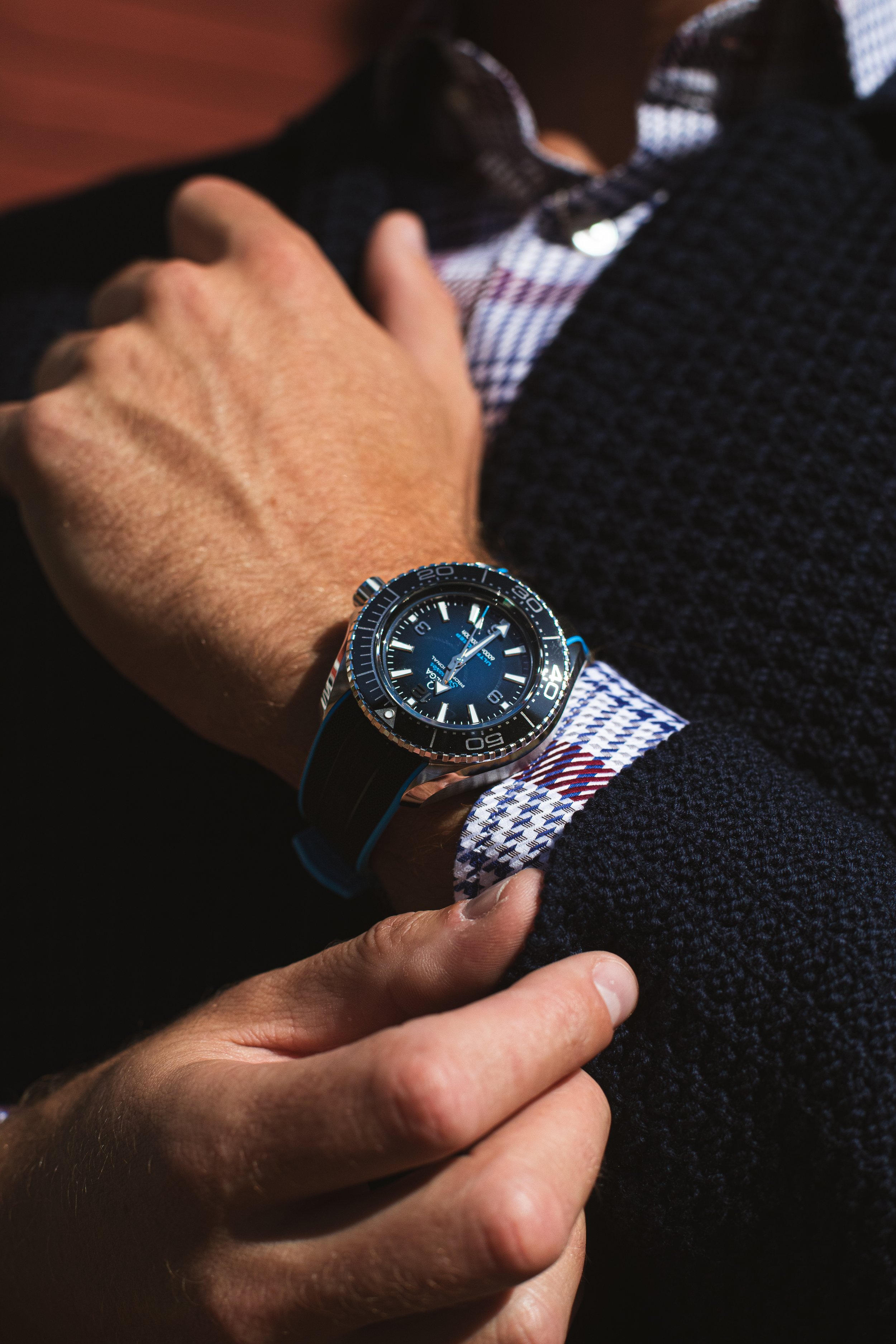 Close-up of a man's wrist wearing a blue and black diving watch, with his hand resting on his chest and wearing a checkered shirt and dark textured sweater.