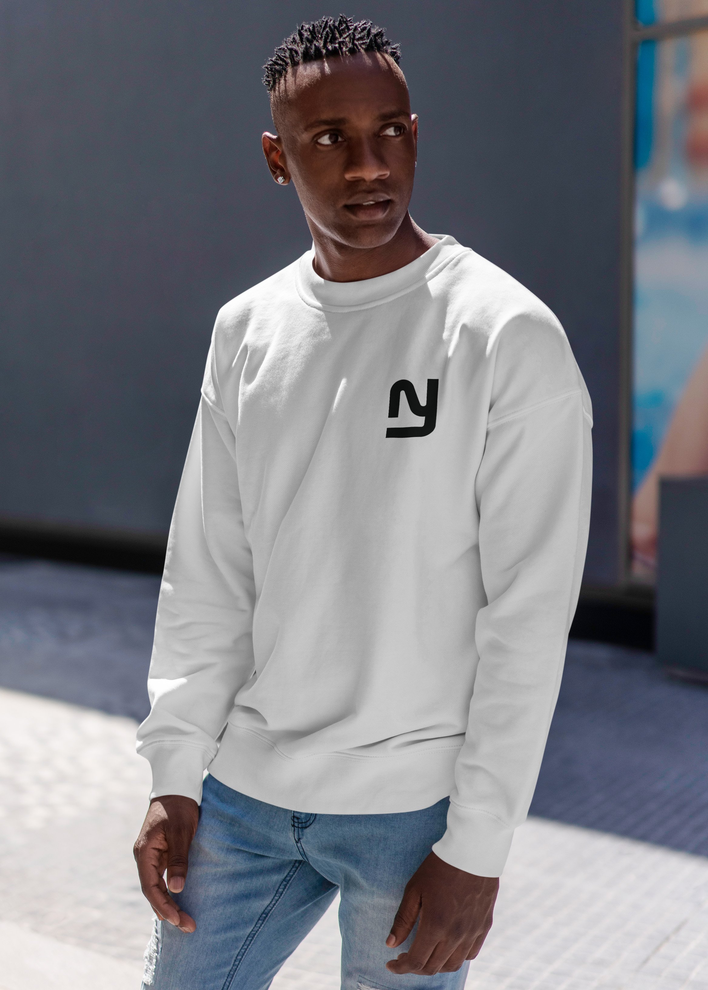 A young man with dark skin and short dreadlocks wearing a white sweatshirt with a black logo on the chest, standing outdoors on a sunny day.
