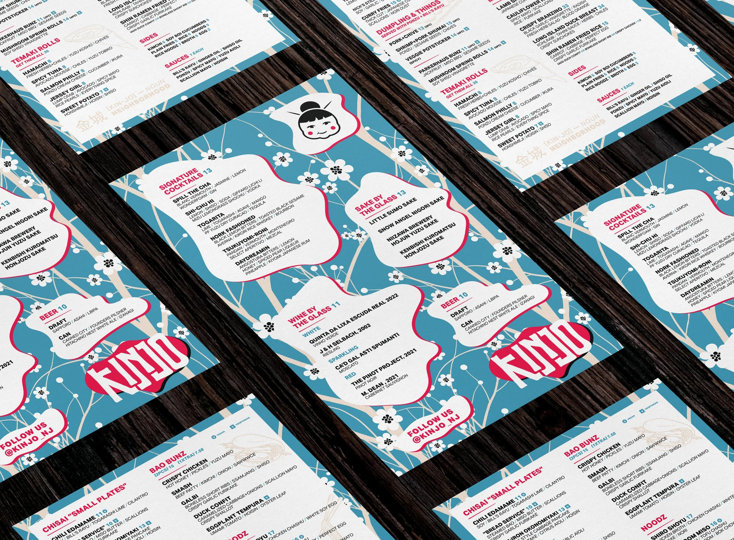Three menus with a blue background, white cherry blossom pattern, and black wood table surface. The menus feature Japanese-inspired illustrations, including a cartoon girl and cherry blossoms, with sections for signature cocktails, sake, wine, and be