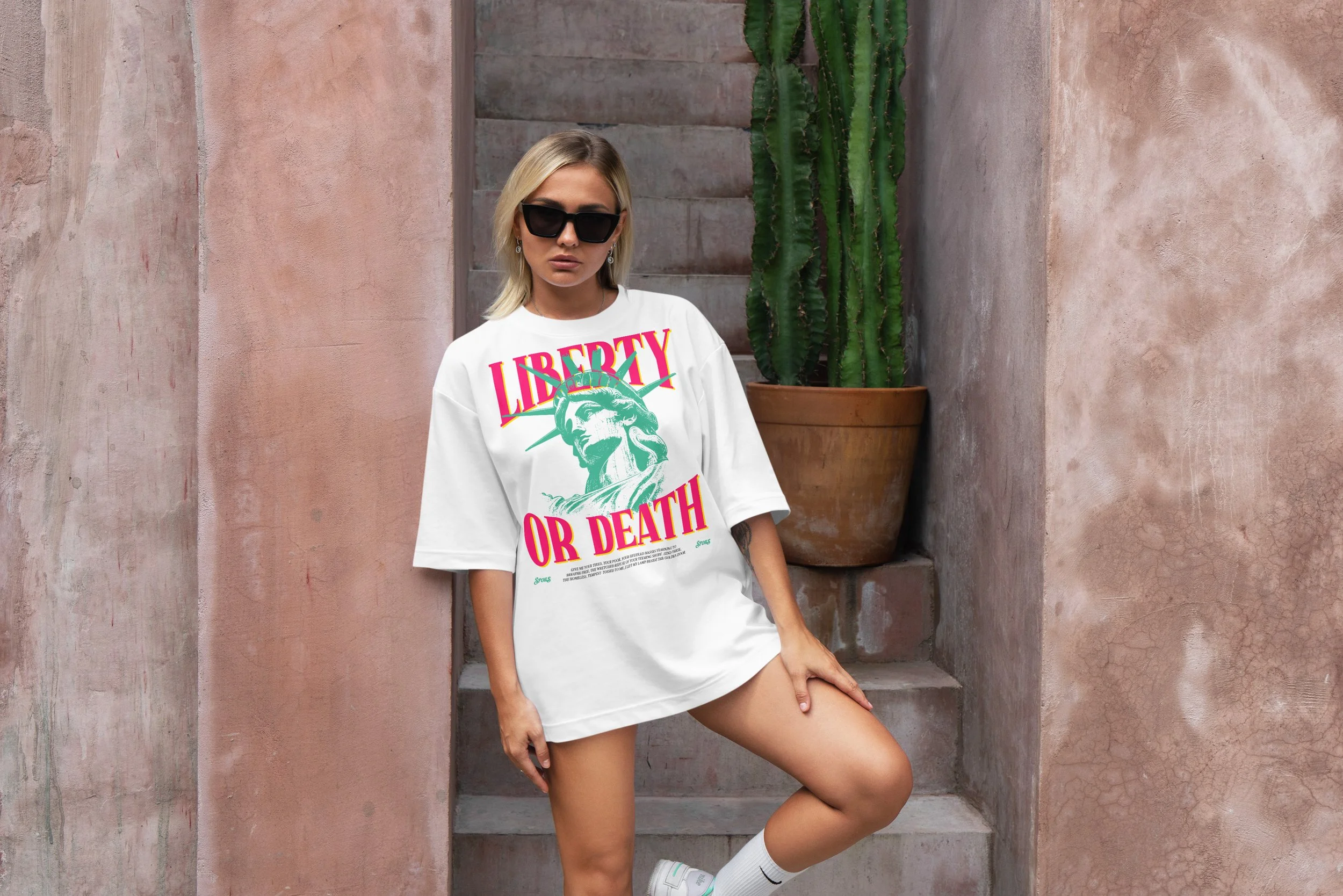 A woman with blonde hair wearing black sunglasses, a white oversized T-shirt with pink and green text and graphic, and white sneakers standing next to a terracotta pot with a tall cactus against a pink textured wall.