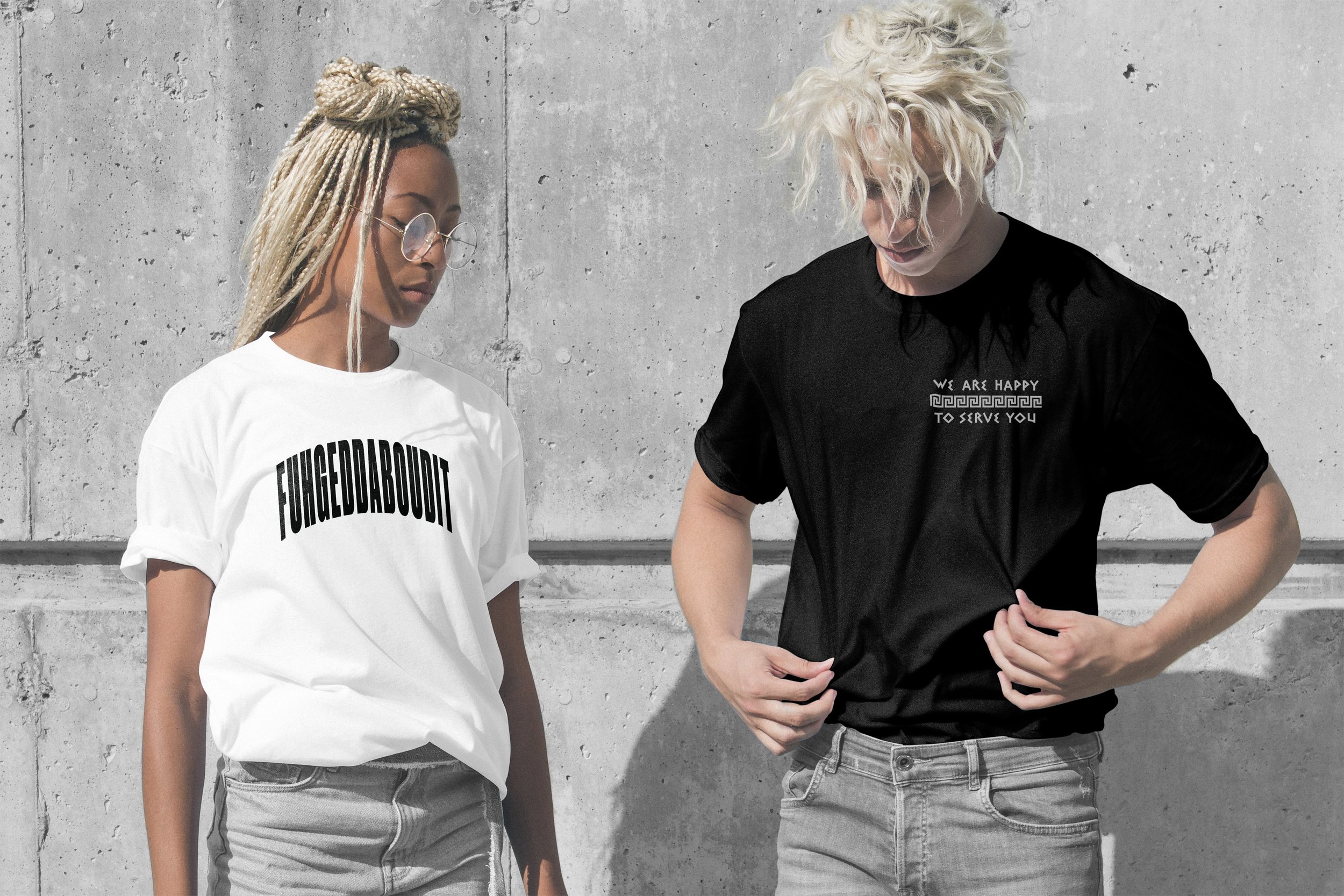 A woman with blonde dreadlocks and glasses wearing a white T-shirt with the text 'FUNGEDABOUDIT', and a man with curly blonde hair wearing a black T-shirt with the text 'WE ARE HAPPY TO SERVE YOU' standing against a concrete wall.