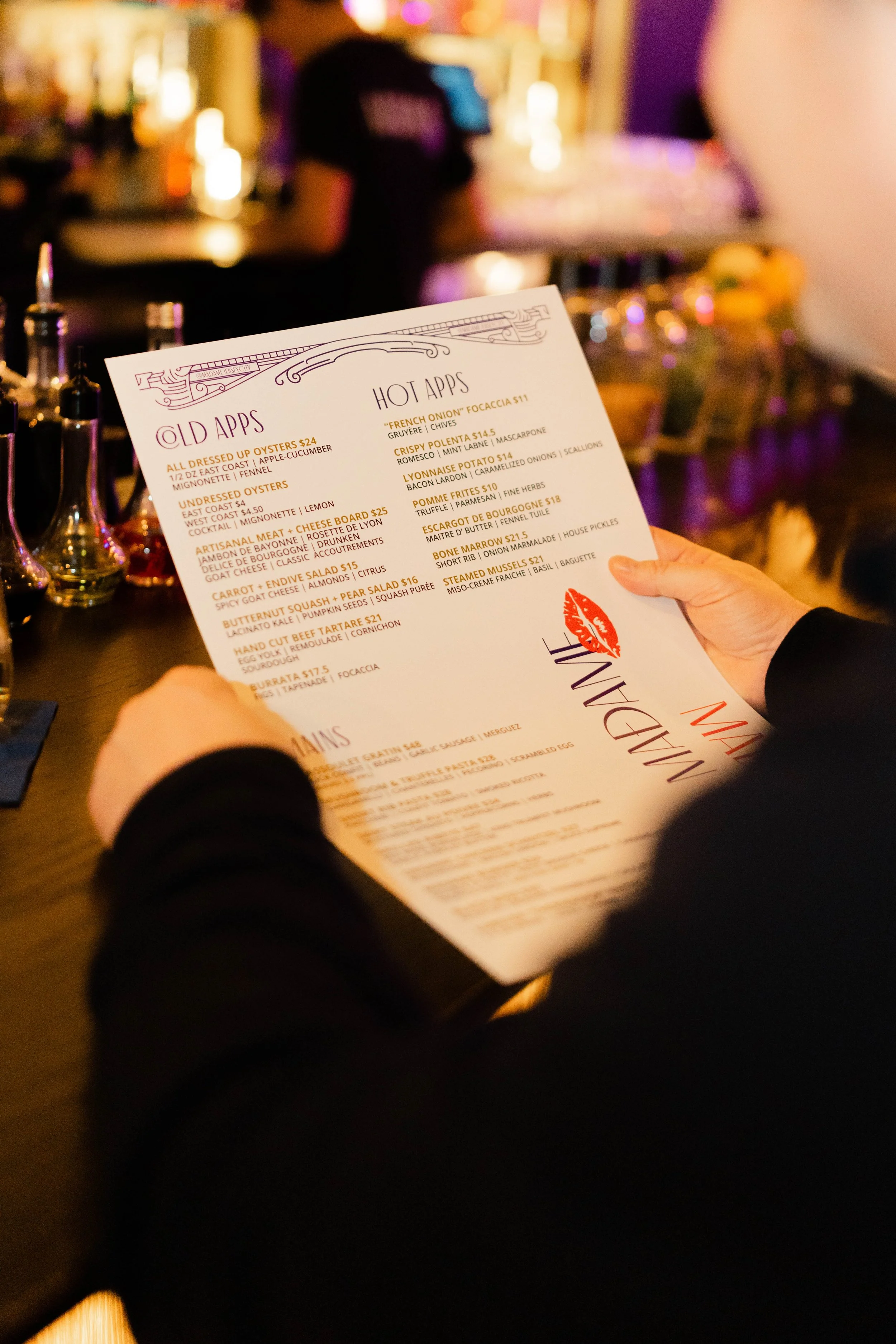 Person holding a menu at a restaurant or bar, with a blurred background of bottles and colorful lighting.