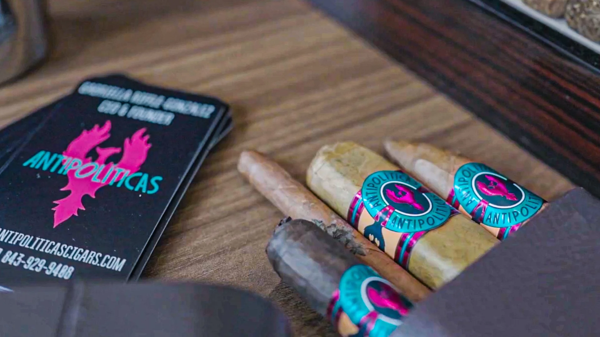 A collection of cigars with colorful bands labeled 'ANTIPOLITICAS' on a wooden surface, next to a stack of business cards for 'ANTIPOLITICAS CIGARS' with pink and blue logo.