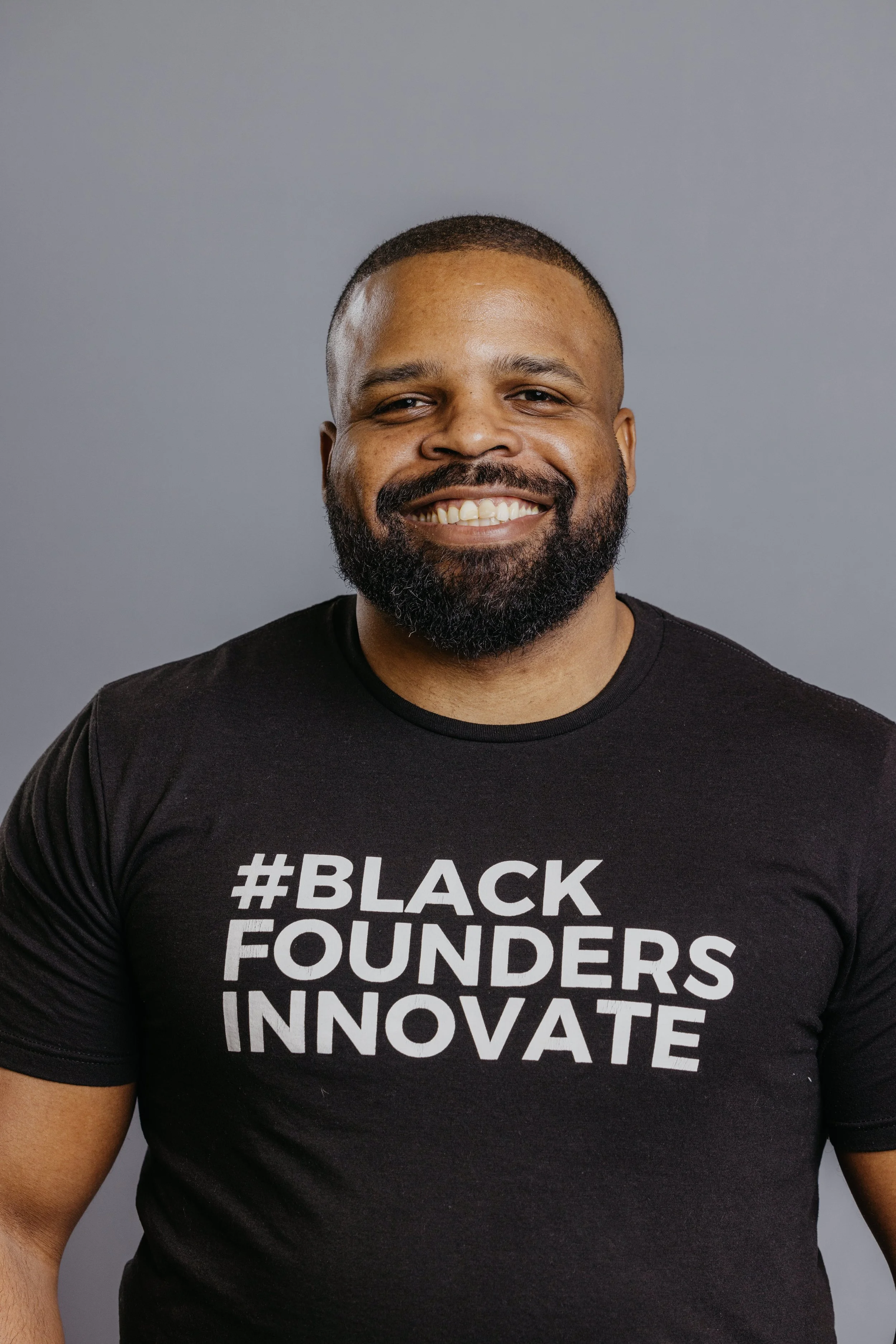 A smiling man with a beard wearing a black T-shirt that says '#BLACK FOUNDERS INNOVATE' stands against a gray background.