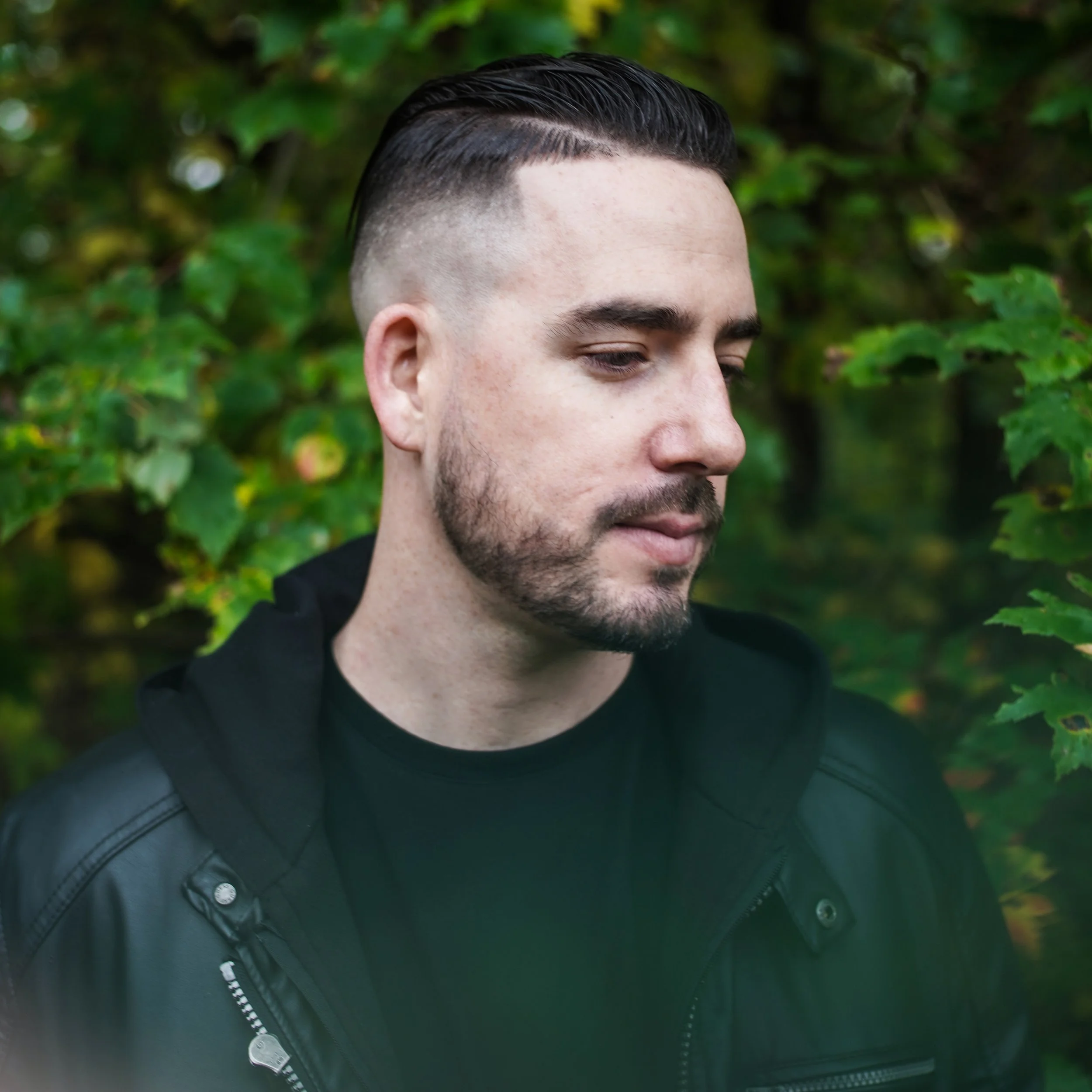 A man with dark wet hair, a beard, and light skin standing outdoors among green leaves, wearing a black jacket and shirt, with eyes closed and a serene expression.