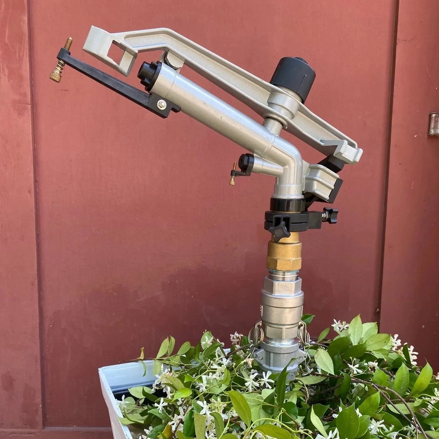 A metal sprinkler head mounted on a bracket, with green leaves and small white flowers in a white container at its base, against a reddish-brown wall.