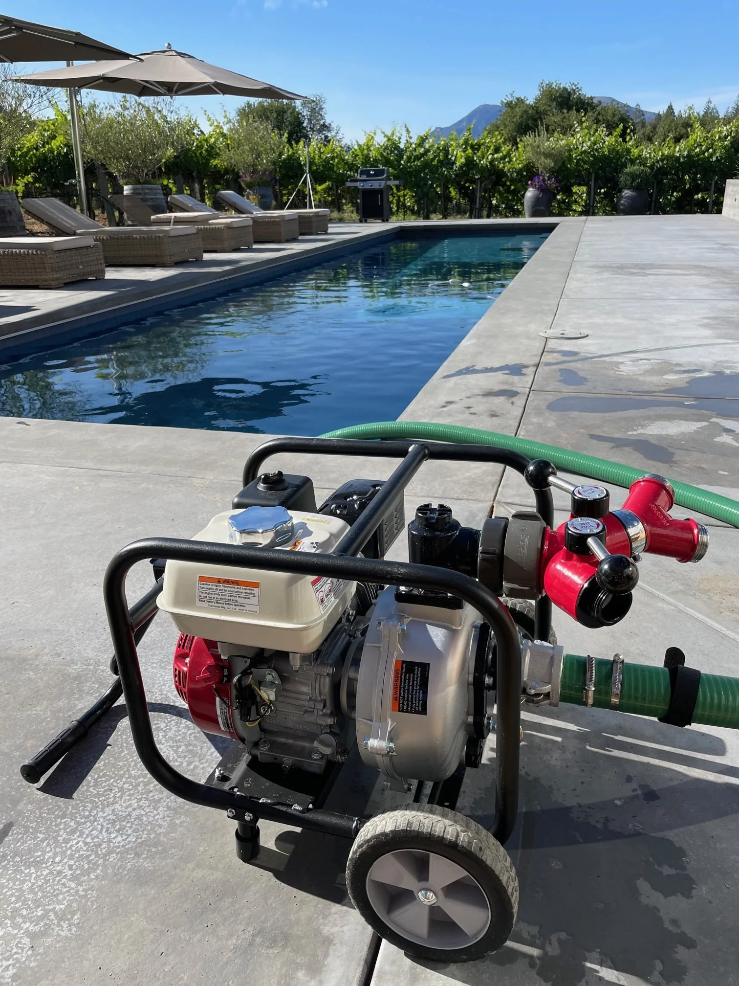 A gas-powered pool pump on a concrete patio beside a swimming pool, with lounge chairs and an umbrella in the background, and trees and mountains in the distance on a clear sunny day.