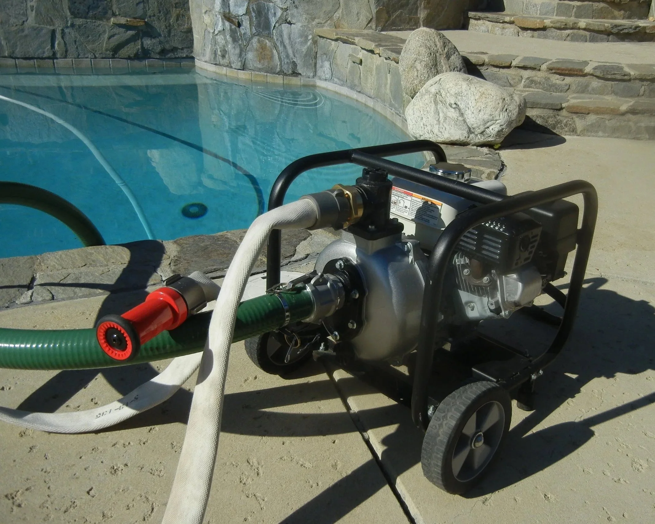 A portable pool pump with hoses connected, positioned near a swimming pool with a stone wall and rocks in the background.