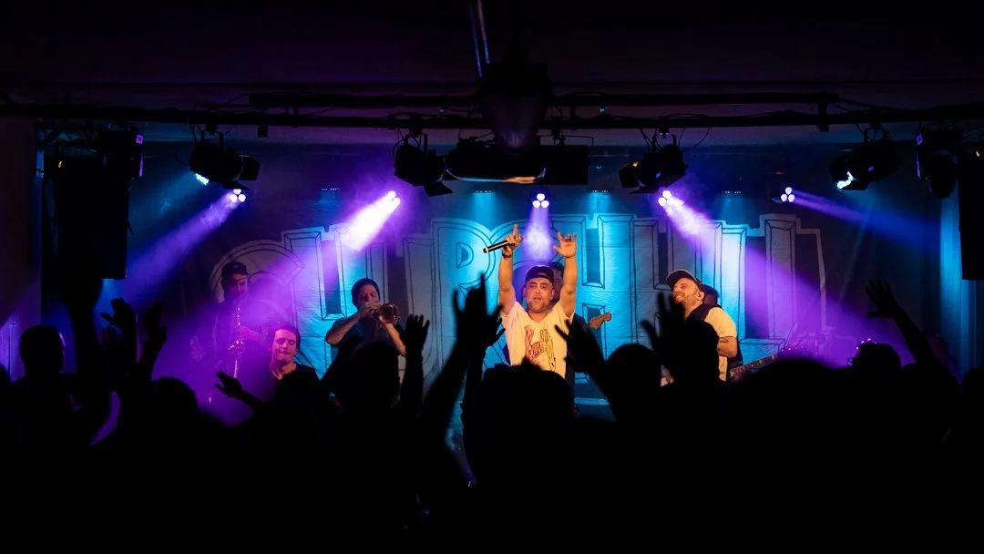 Ein Konzertbild von einer Band die auf der Bühne steht. Das Publikum streckt die Hände in die Luft