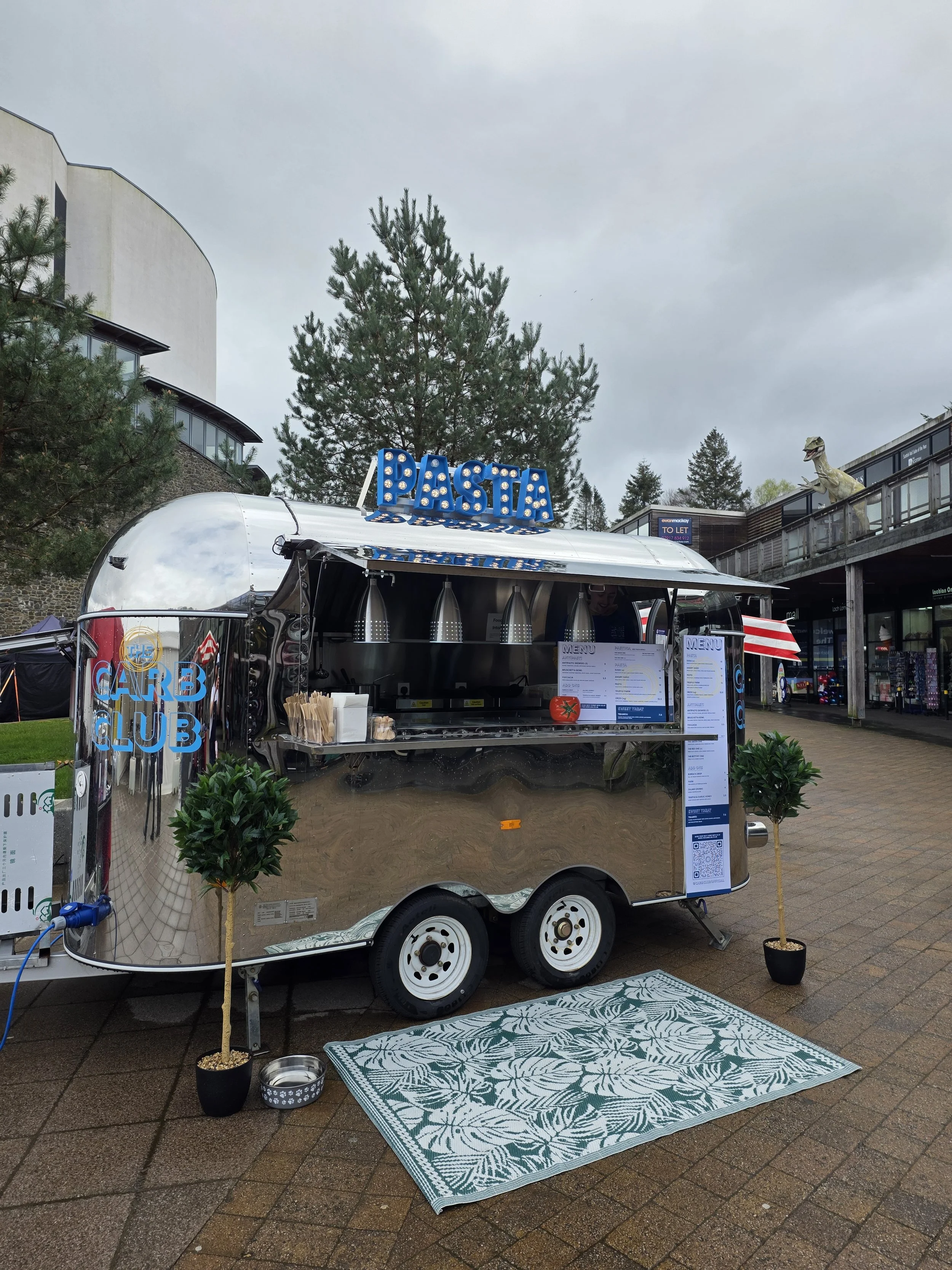A food truck labeled 'Pasta' with a reflective surface, parked outdoors on a cloudy day, with a decorative rug, two potted plants, and dog bowls in front. There is a large dinosaur statue and retail stores in the background.