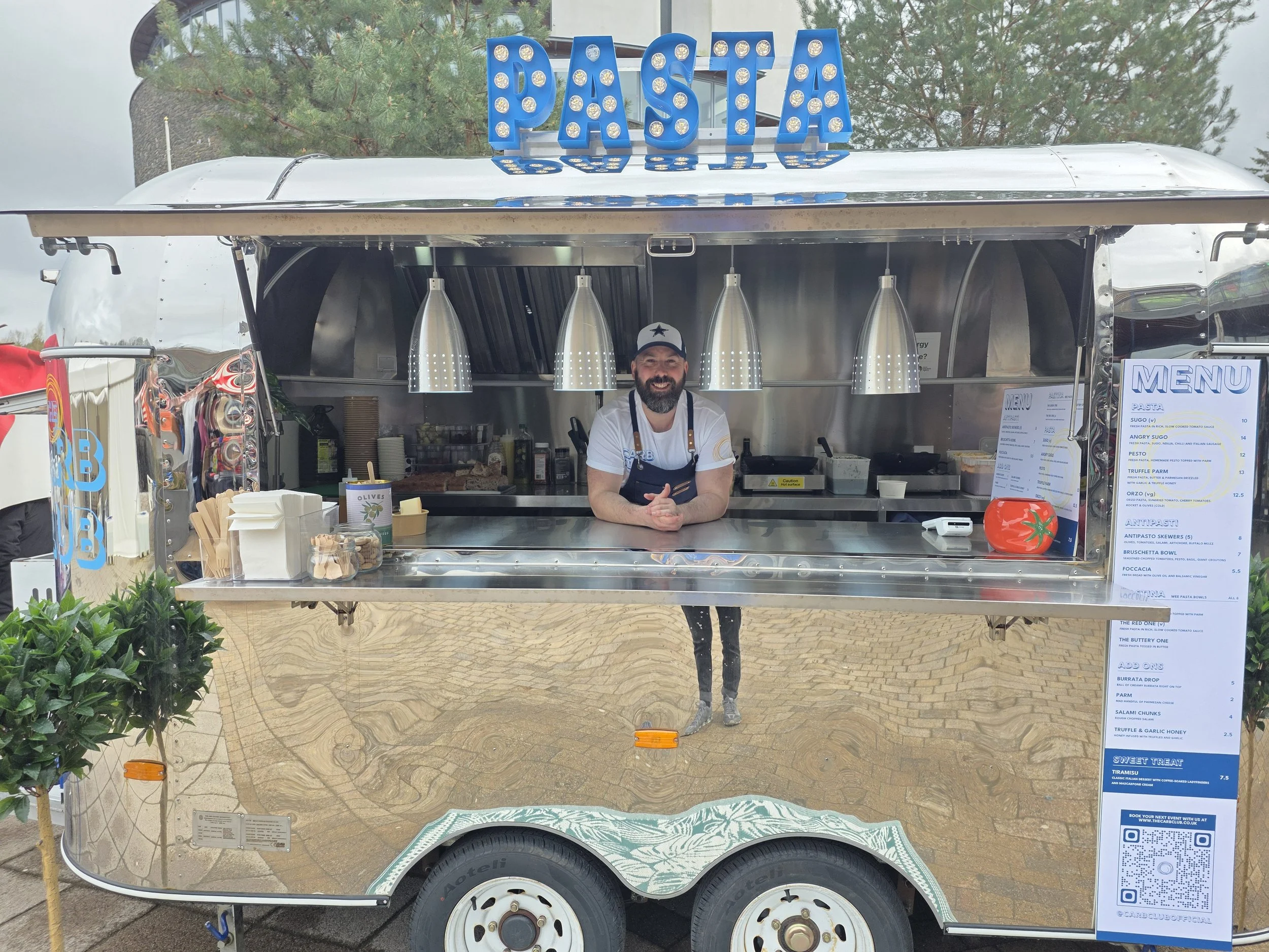 A food truck with a large blue sign that reads 'PASTA' on top. The truck has a man with a beard and a Star Wars cap smiling and standing at the counter. The menu is visible on the side, listing different pasta dishes. The truck is parked outdoors, with trees in the background.