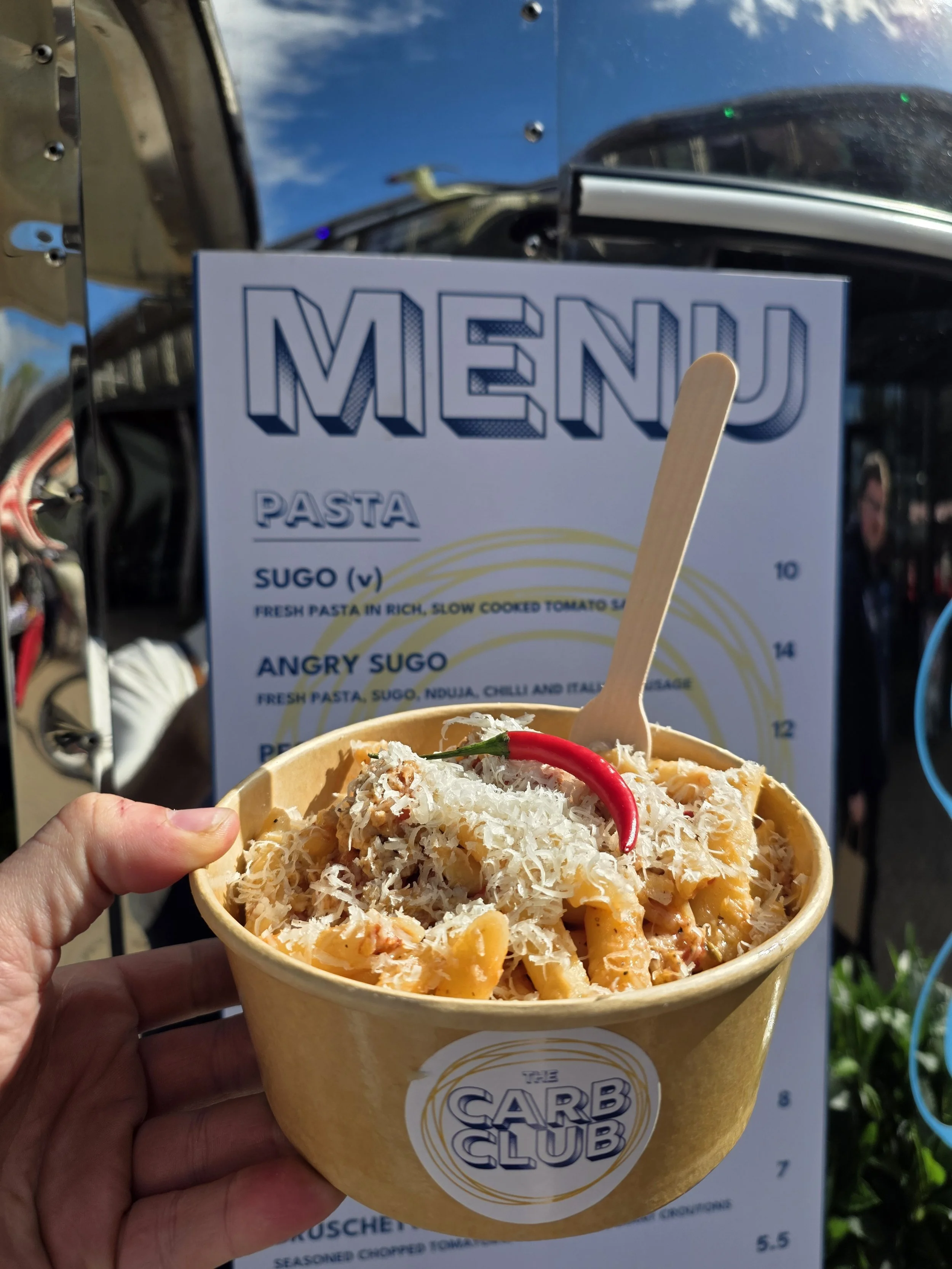 A hand holding a cup of pasta with grated cheese and a red chili pepper on top, in front of a menu from The Carb Club, outdoors on a sunny day.
