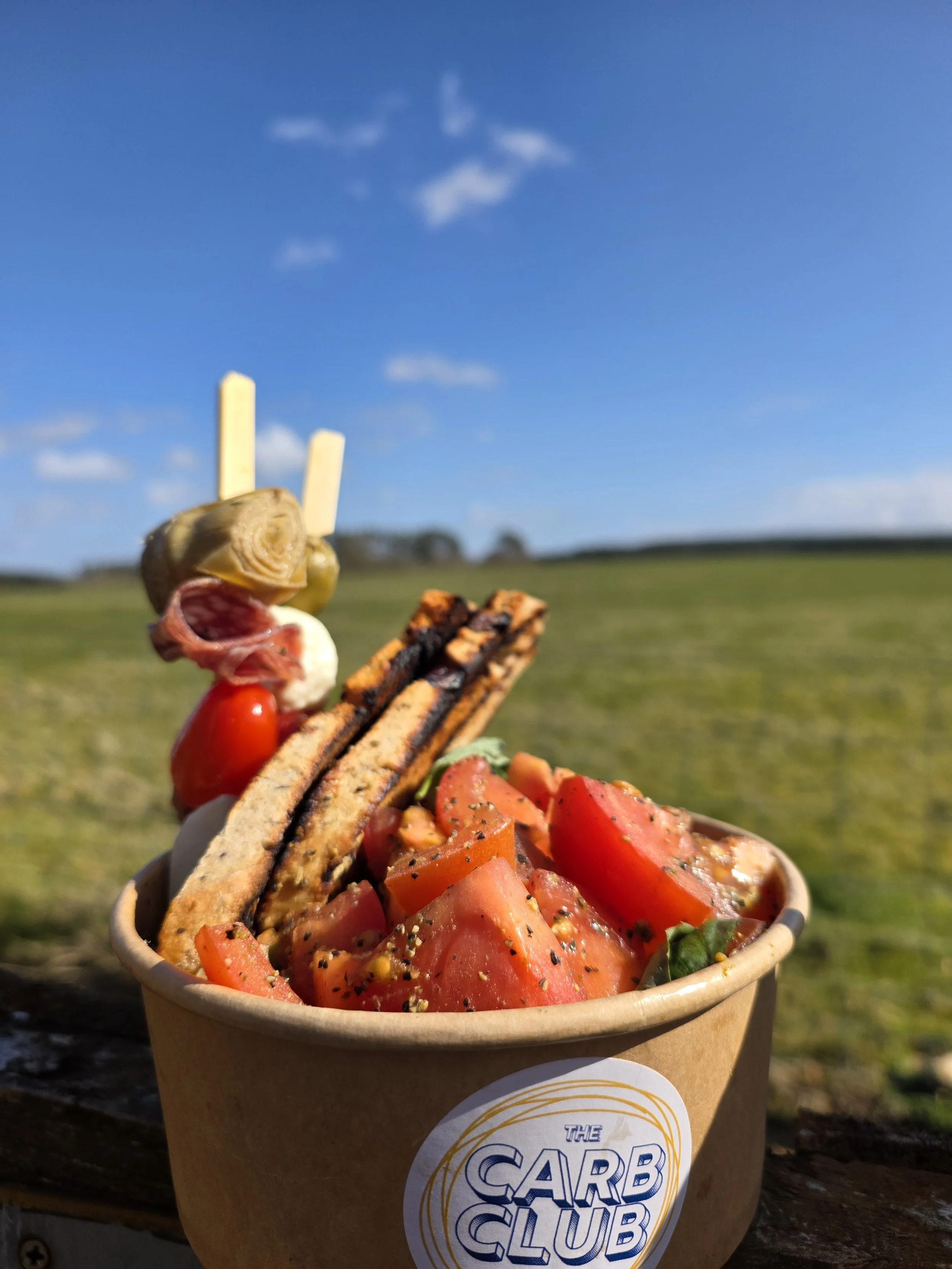 A bowl of chopped tomatoes, grilled vegetables, and slices of bread with toppings, outdoors under a blue sky with clouds.
