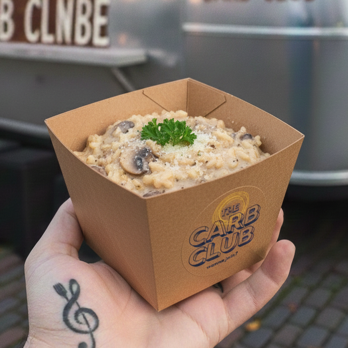 A hand holding a square brown paper box of creamy pasta with black olives, garnished with a parsley sprig.