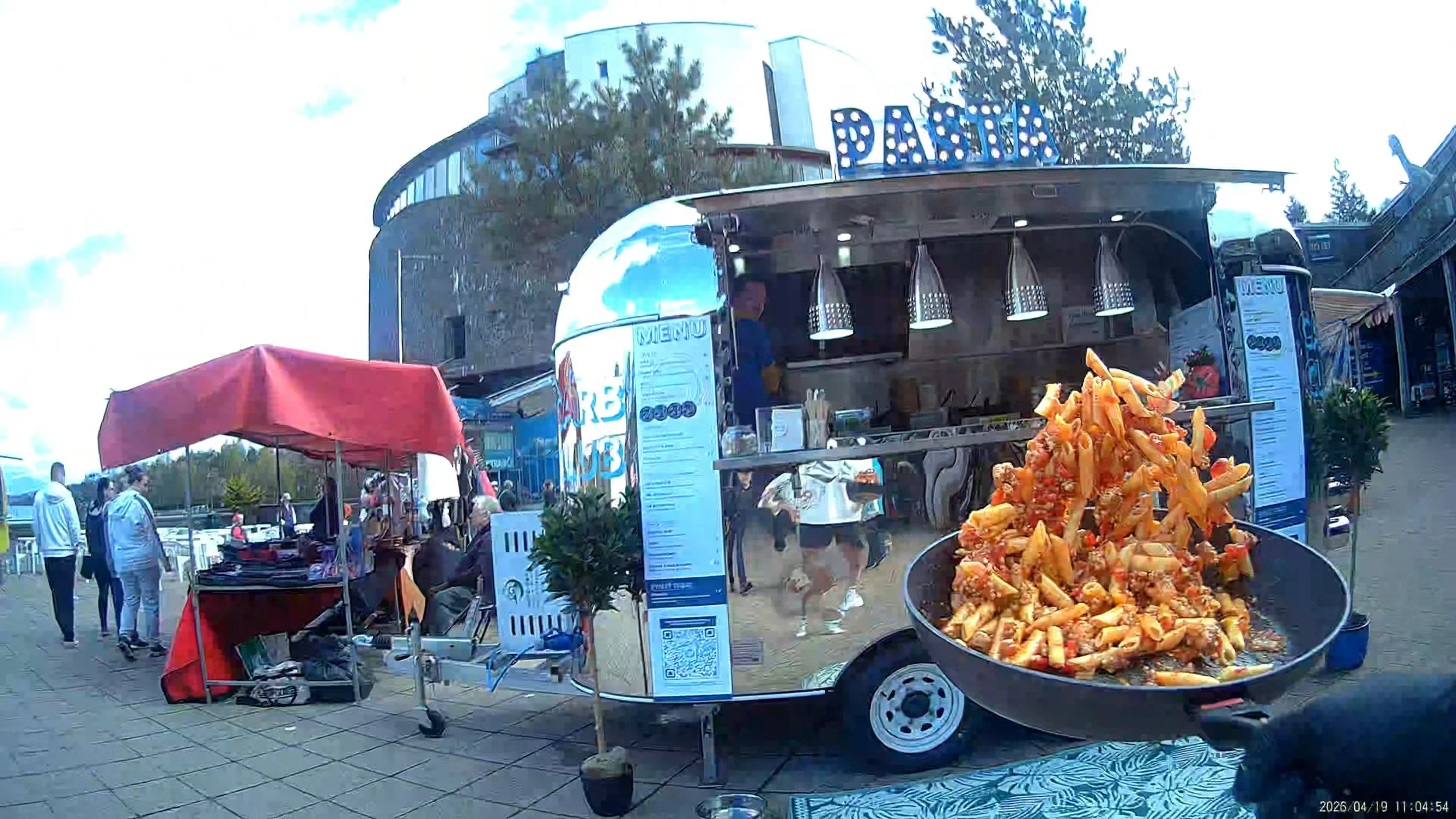 Food truck with large sign at the top spelling 'DASIA.' A display of orange food items, possibly chicken wings, in a round container in the foreground. People walking and browsing in an outdoor market area; red tent on the left and multiple pop-up stands. A building is in the background with some trees and a partly cloudy sky.