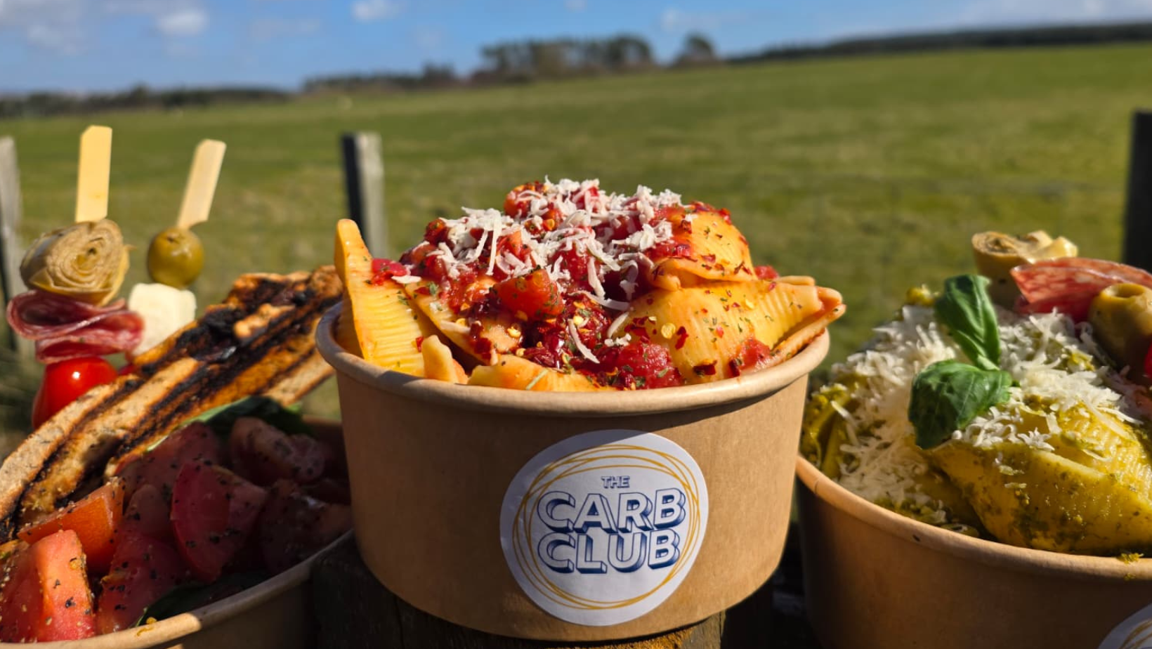 Bowl of pasta topped with cheese, tomato sauce, and herbs, with a label reading 'The Carb Club', outdoors with green grass and blue sky in the background.
