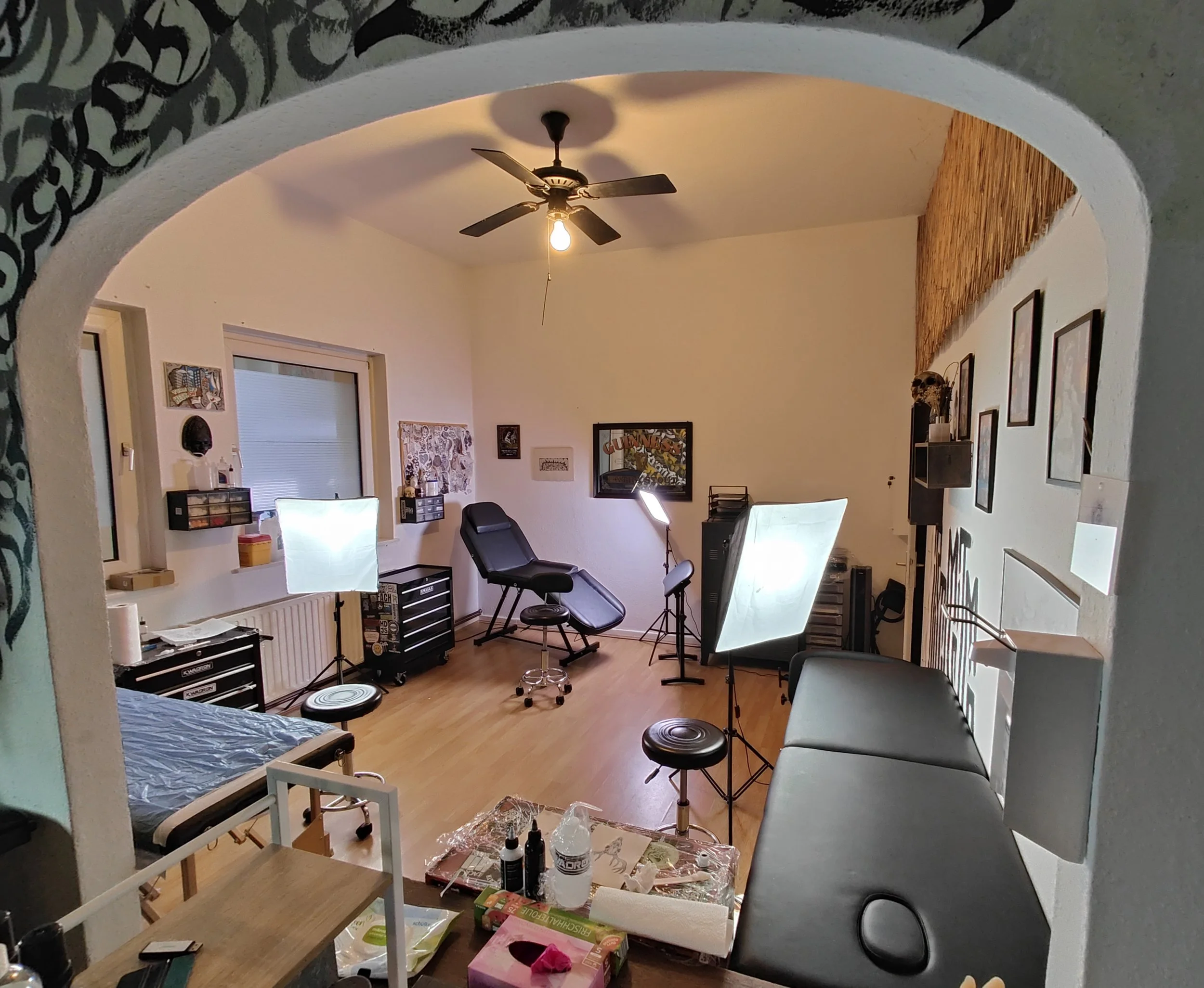 Ein Tattoo-Studio mit  Untersuchungsliegen, Fotostudio- und Tattooausrüstung mit mehreren Stühlen, Blick durch einen Bogen in die Szene. Es gibt eine Deckenlampe, einen Ventilator, einen Schreibtisch, Bilder an der Wand.