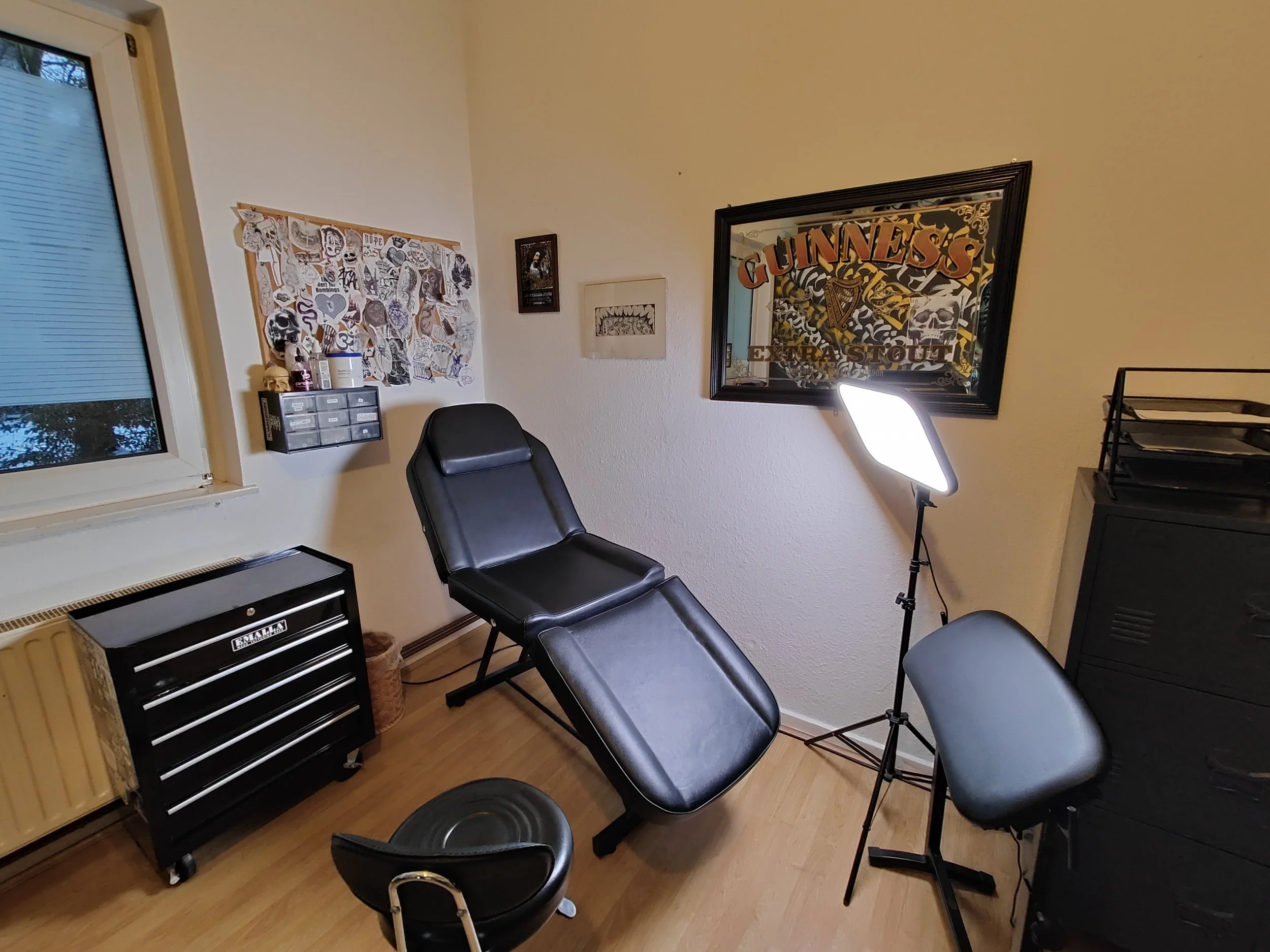 Tattooarbeitsplatz mit Tattooliege, schwarzer Sitzhocker, Stehlampe, Wandbilder, Fenster, Dunkler Schrank, kleine Aufbewahrungsboxen, Pinnwand mit wannadoos oder tattoo stencil.