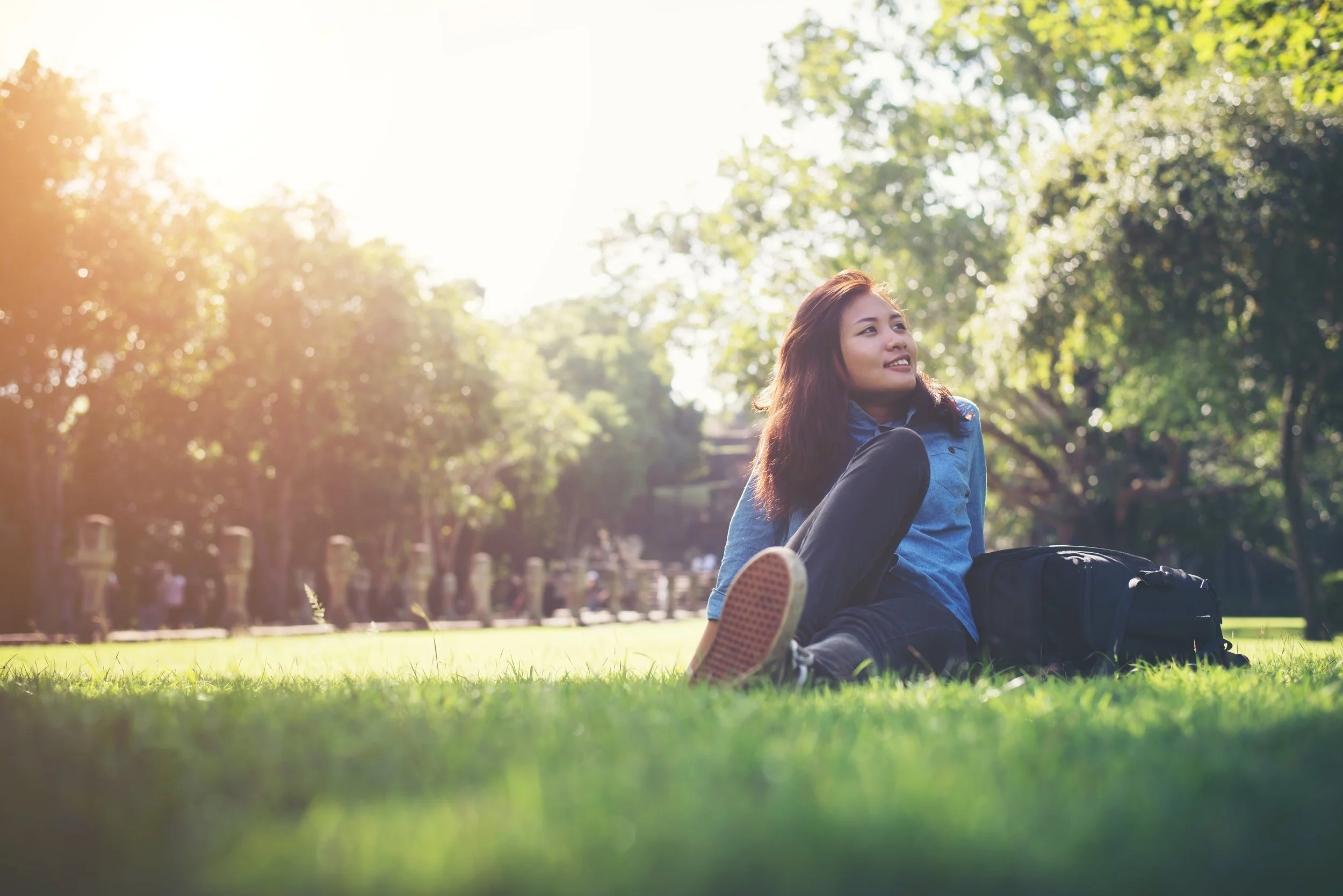 A young woman sitting on the grass in a park during daytime, with trees and sunlight in the background.