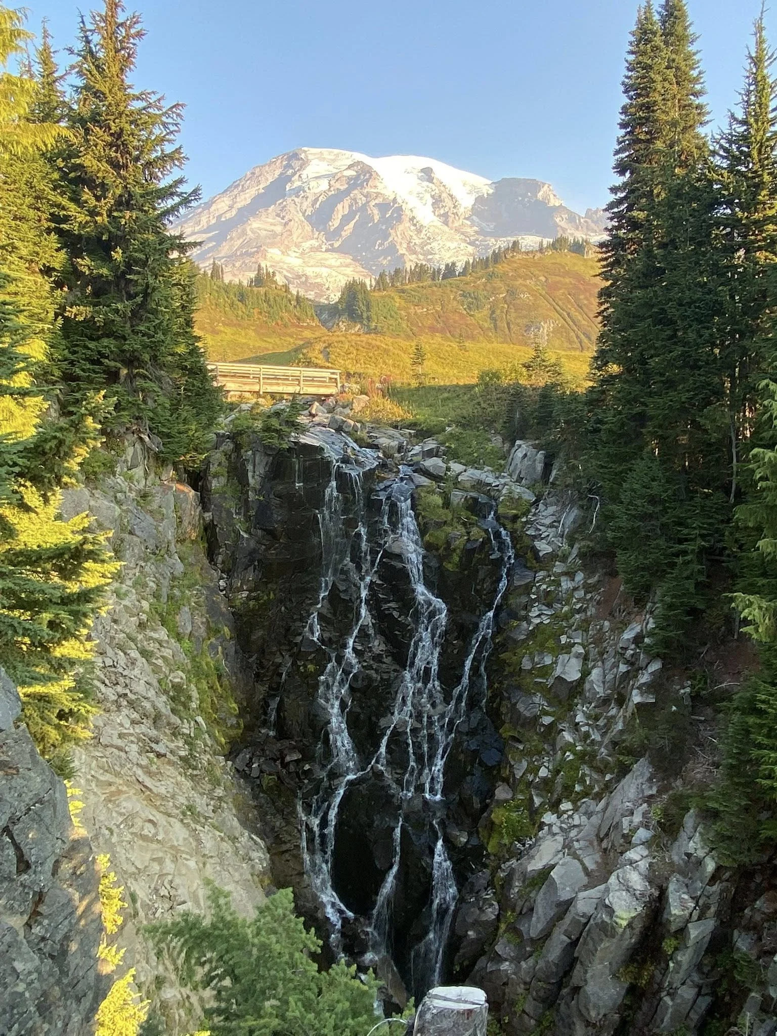 Snow-capped mountain with a waterfall cascading down rocky cliffs surrounded by greenery and tall trees. nature for mental health, emotional well being, nature walk, forest bathing, exercise, lifestyle modifications, wellness, caring minds llc