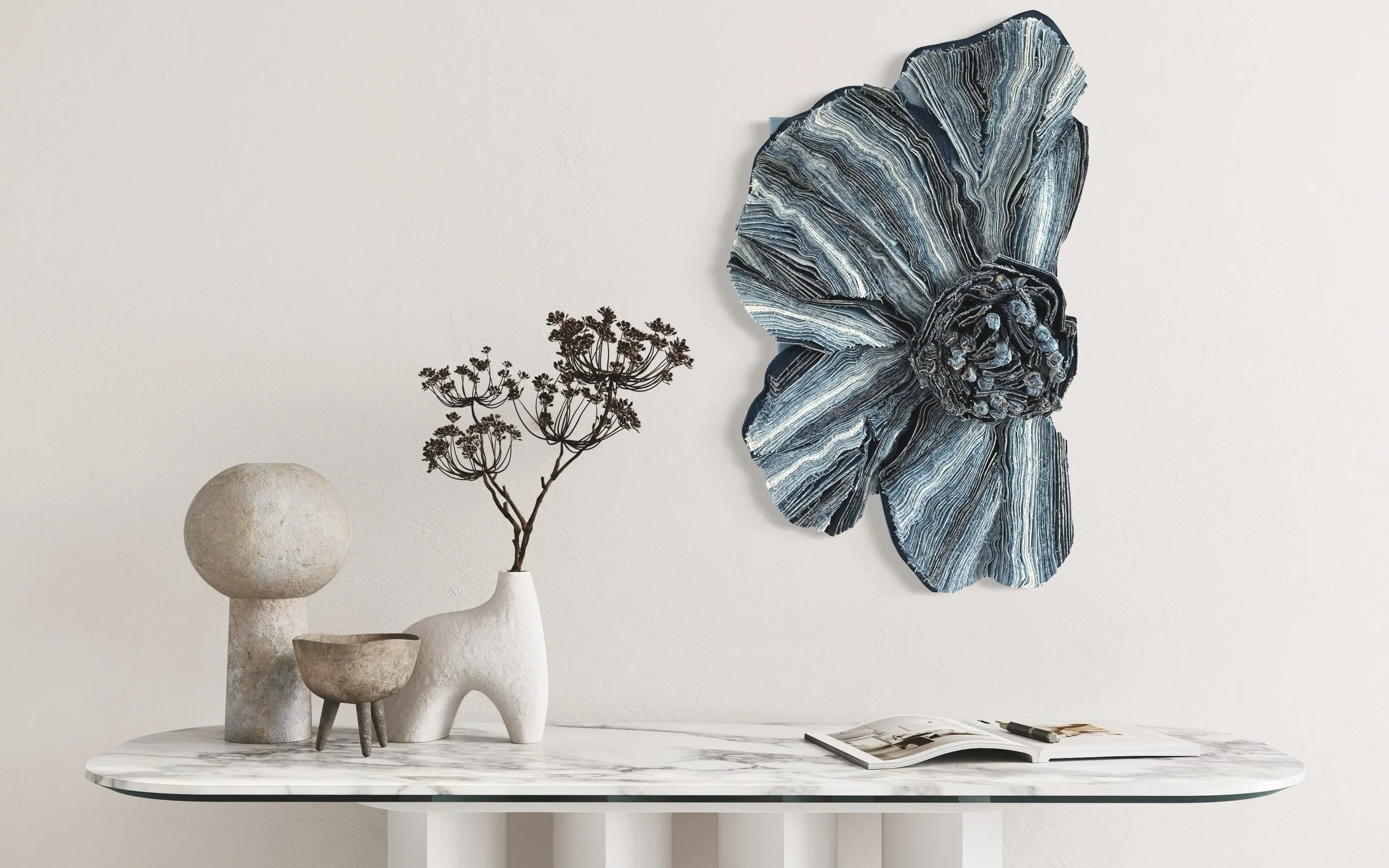 Decoratieve kunstwerken en objecten op een witte marmeren tafel, waaronder een keramieken vaas met een tak, een klein gestileerd beeld, en een boek, met een grote blauwe bloem wanddecoratie op een witte muur op de achtergrond.