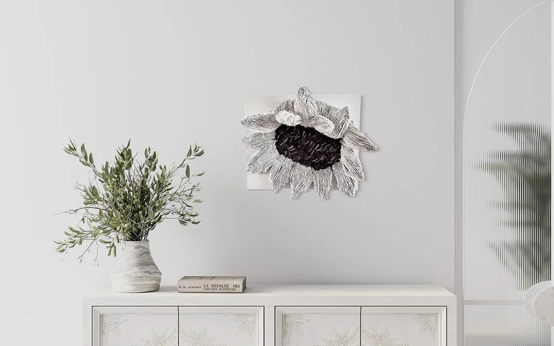 Een minimalistische interieurafbeelding met een witte muur, een decoratieve textiele muurkunst van een grote zonnebloem, een witte vaas met groene bladeren en een boek op een witte kist.