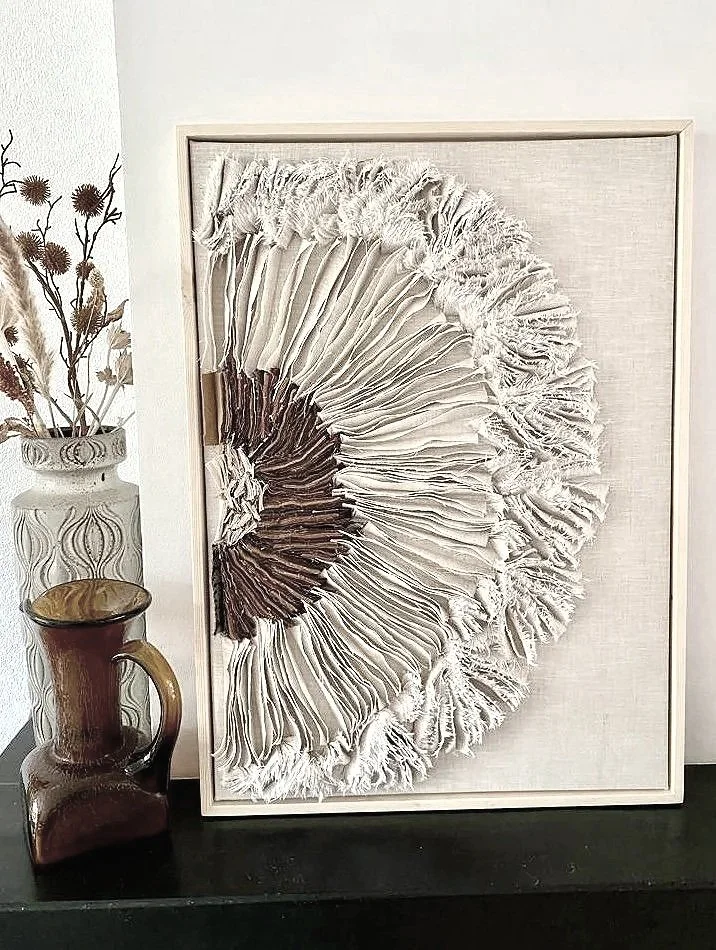 Een decoratief kunstwerk met een driedimensionaal ontwerp dat lijkt op een paddenstoel of een bloem, met gerstekorrel-achtige textuur in beige en bruine tinten. Het staat op een zwarte tafel naast een vaas met gedroogde bloemen en een kleine bruine v