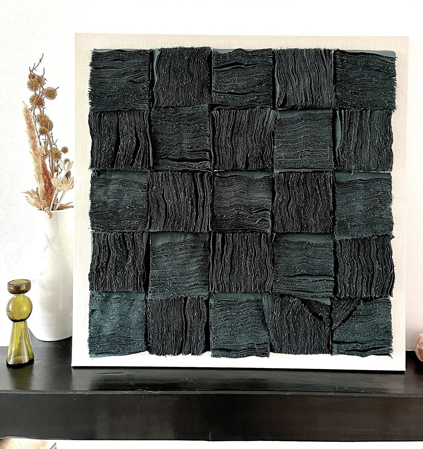 Vierkante sculptuur gemaakt van zwart papier met gestructureerde, golvende lijnen, bevestigd op witte achtergrond, geplaatst op zwarte tafel naast een vaas met gedroogde bloemen en een kleine groene glazen fles.