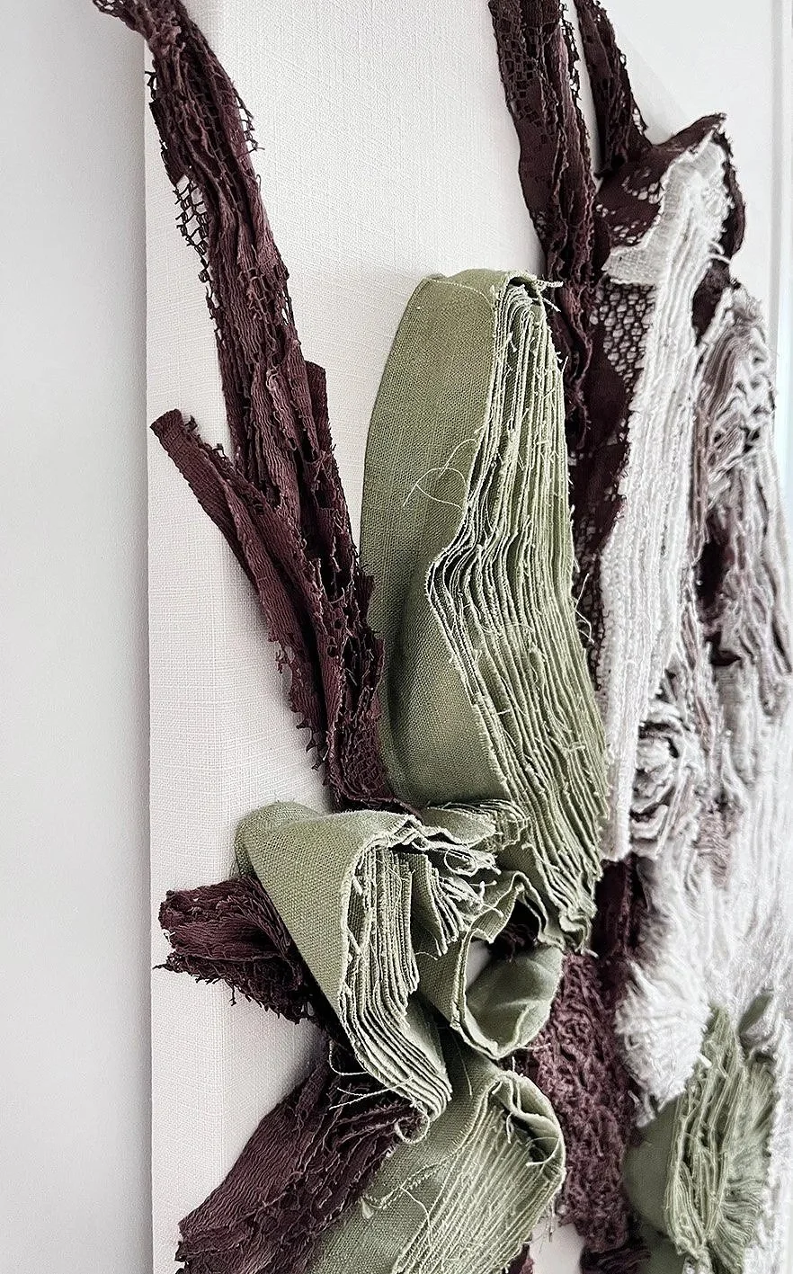 Detail van een textielkunstwerk met gevouwen en gerafelde stoffen in tinten groen, bruin en wit, bevestigd op een witte muur.