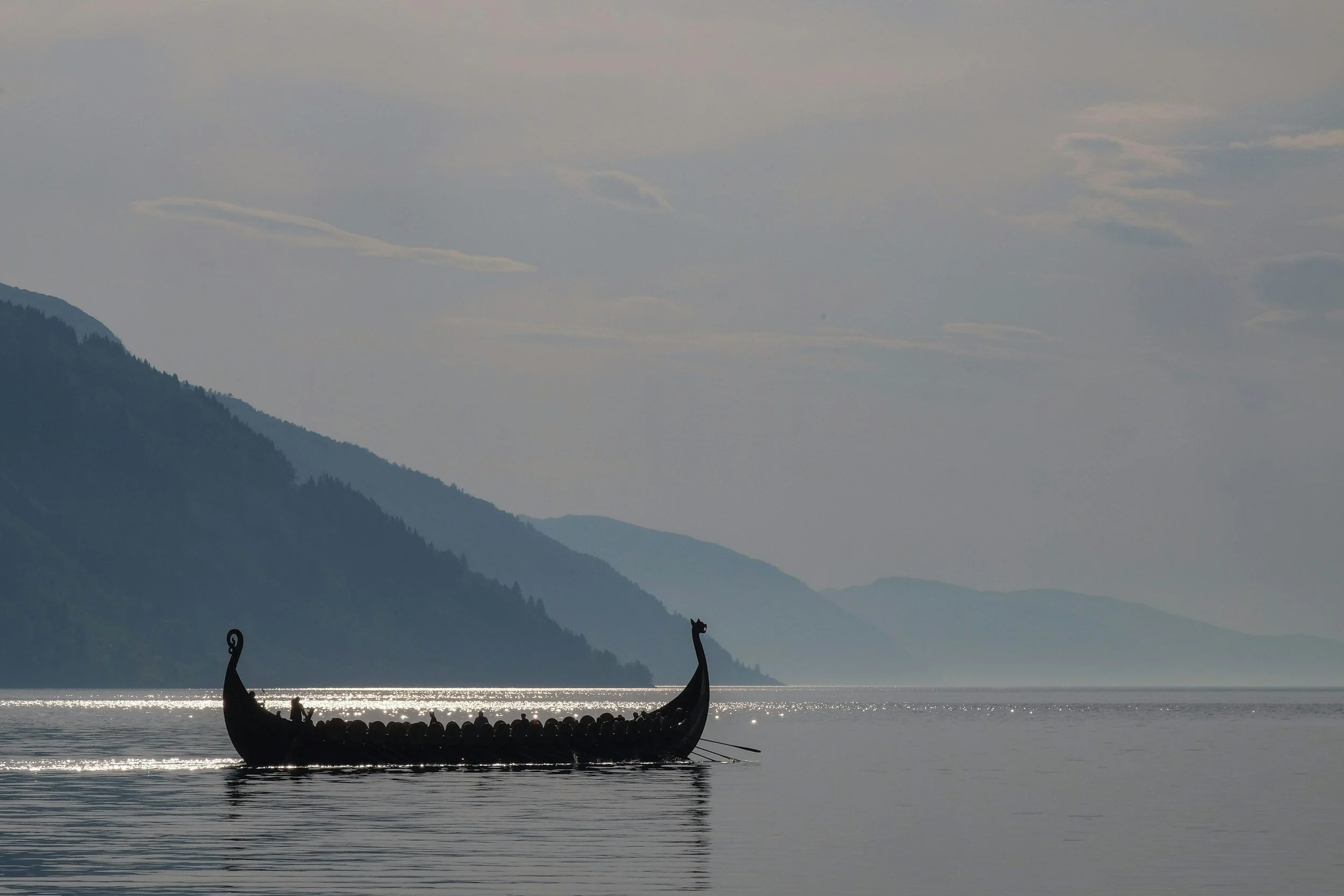 Een silhouet van een houten Vikingboot met drakkars ornamenten op het water, met een berglandschap op de achtergrond en een bewolkte hemel.
