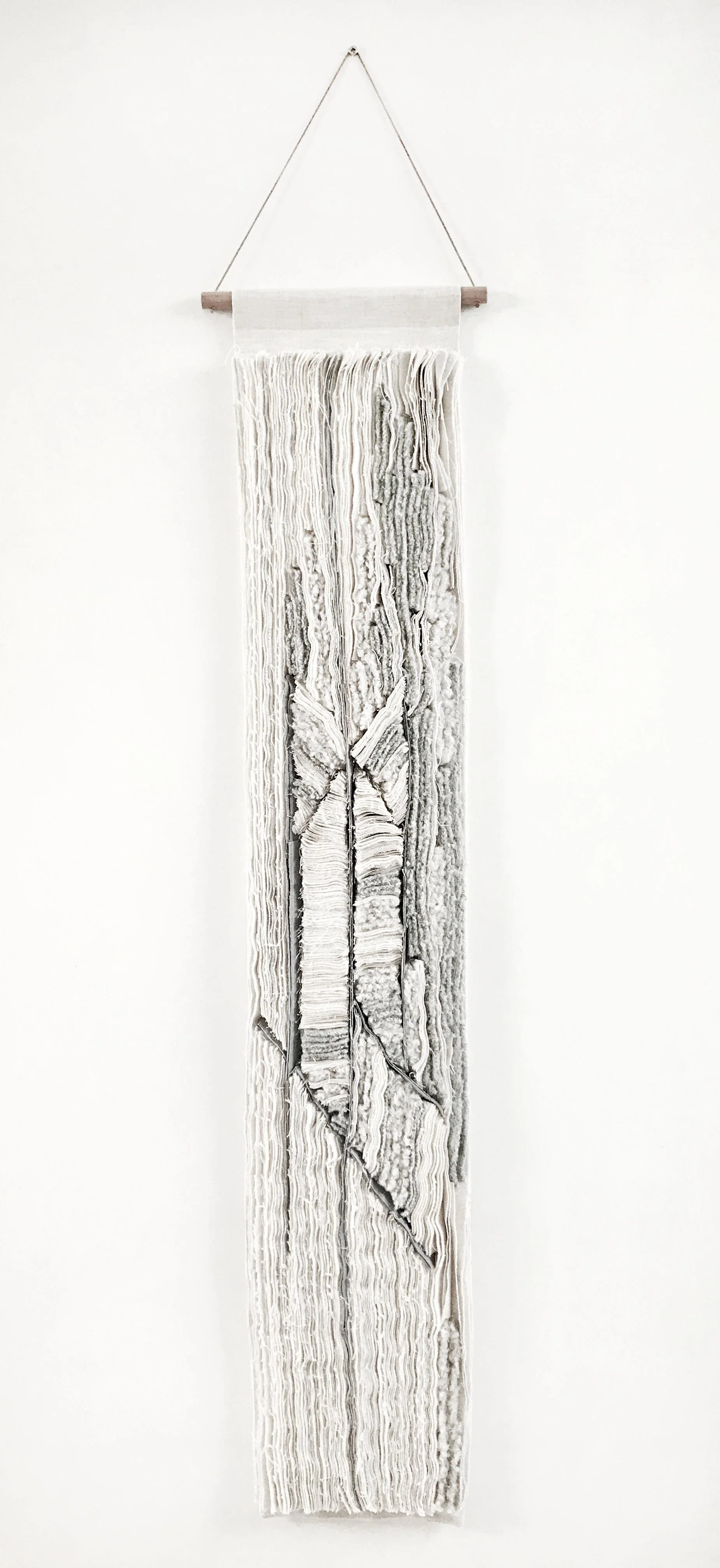 Wit en grijs textielkunstwerk met textuur, hangend aan een houten stok en touw aan een witte muur.