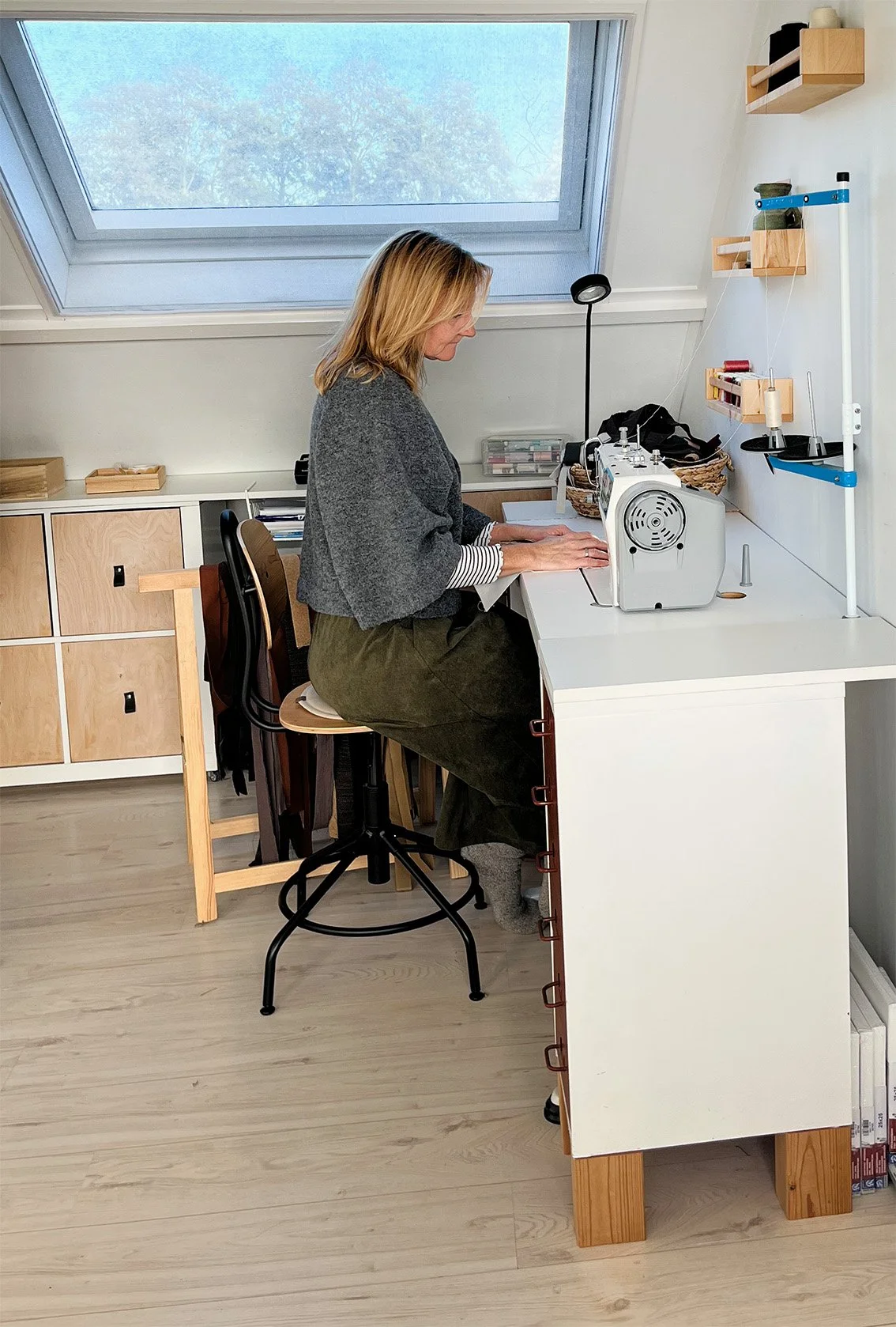 Vrouw die met een naaimachine aan een witte werktafel in een lichte kamer in een schuine dakruimte werkt.
