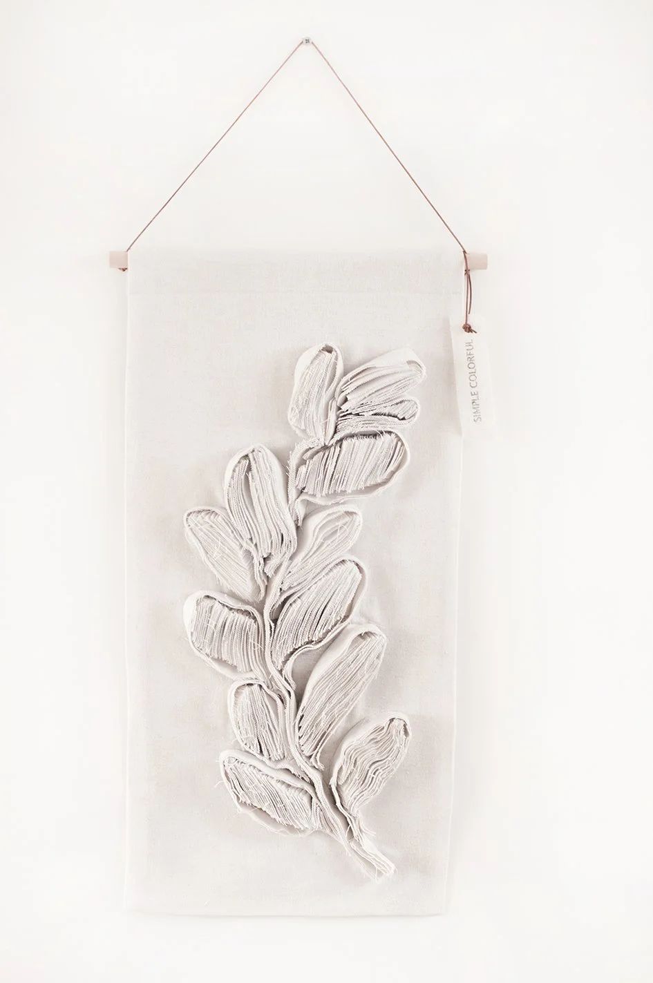 Wit textielkunstwerk met gevouwen stoffen die een blad vormen, bevestigd aan een witte achtergrond en opgehangen aan een dunne draad.