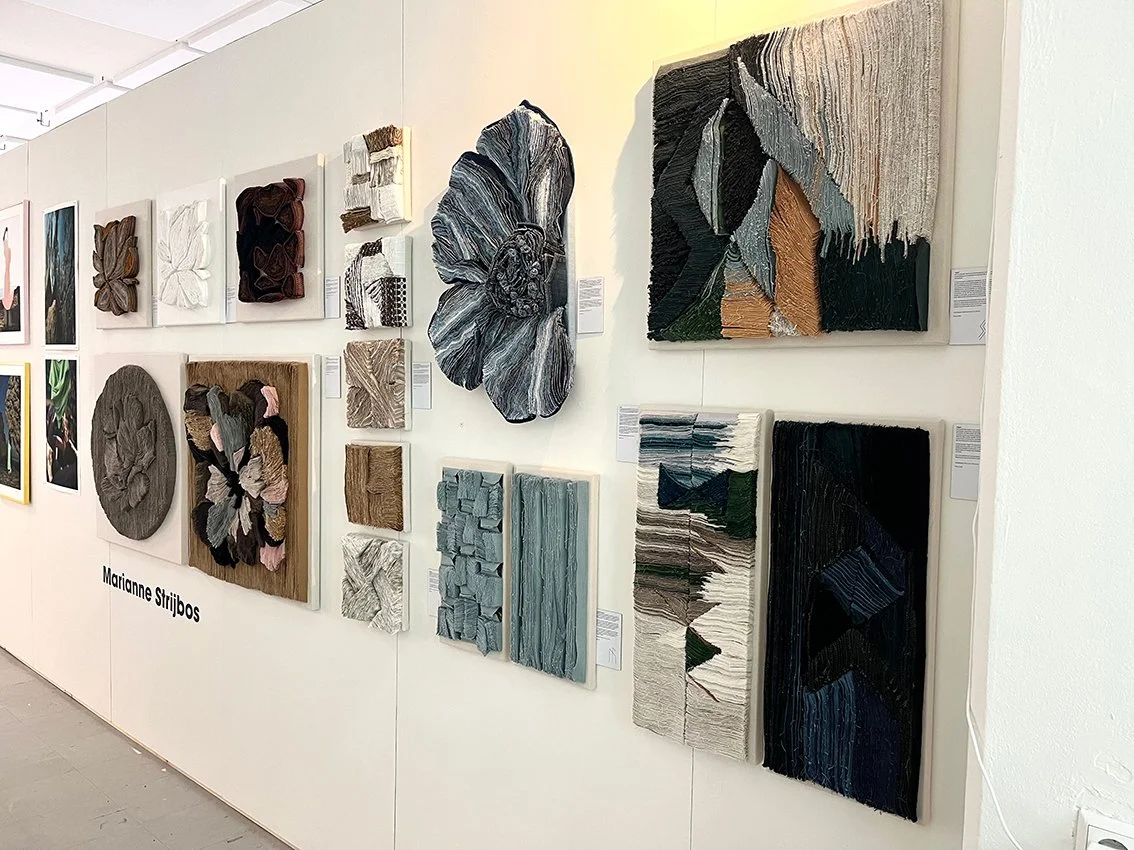 Tentoonstelling van textielkunstwerken op een witte muur, met verschillende abstracte en botanische ontwerpen in verschillende formaten en kleuren.