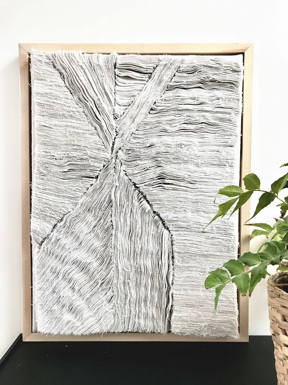  abstract vlakkunstwerk met textuurrijke, witgekleurde lagen in een houten lijst, naast een groene plant in een mand.