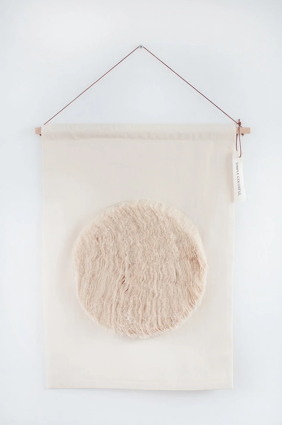 Een muurkunstwerk met een zachte, ronde, beige textuurcirkel op een witte stoffen achtergrond, opgehangen aan een houten stang en een rood koord.
