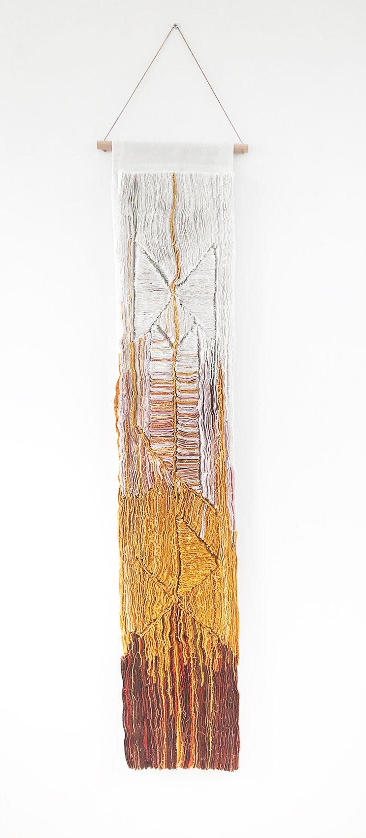 Geweven wanddecoratie met abstract patroon in wit, geel, oranje, rood en bruin op een witte muur.