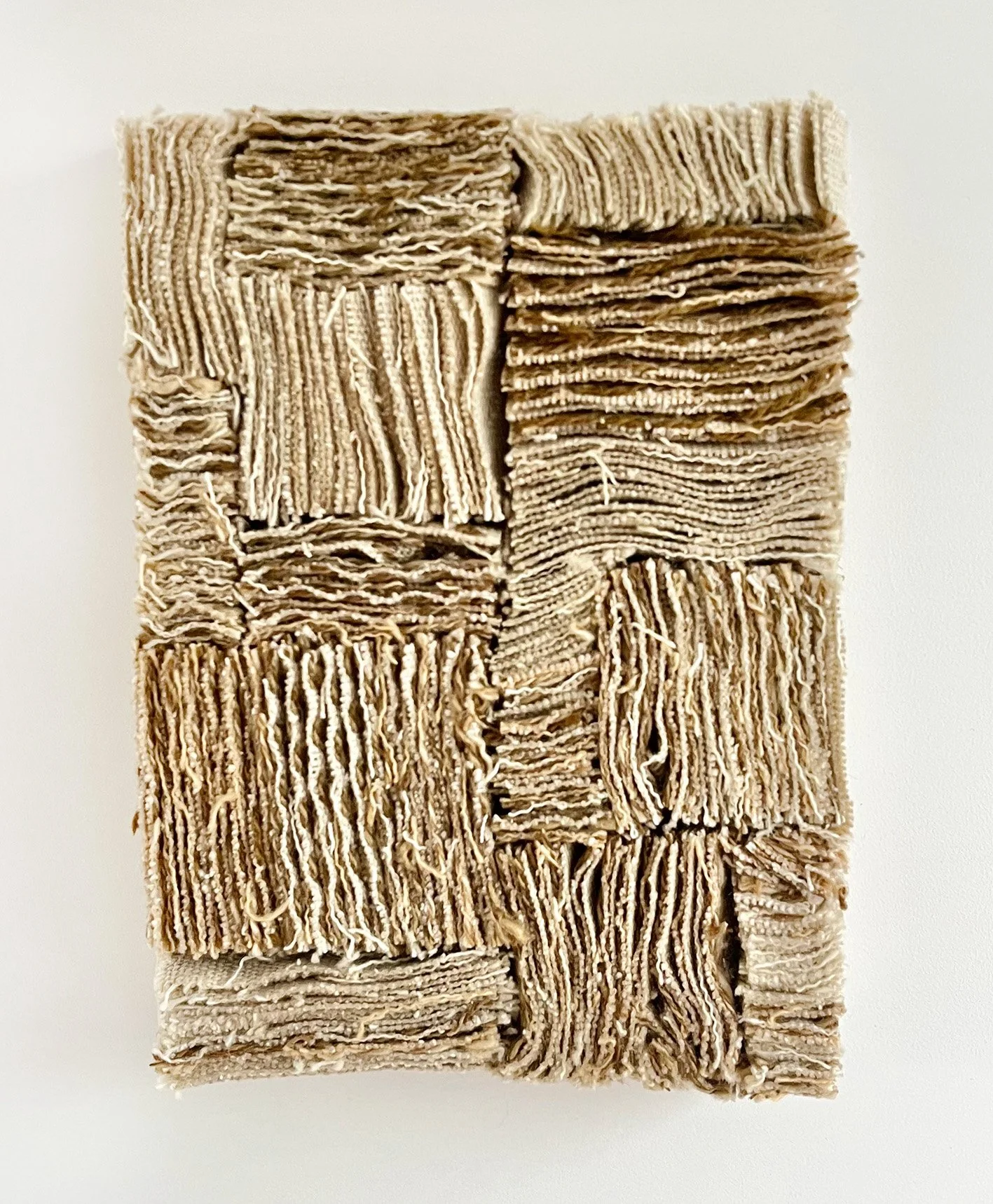 Een kunstwerk van samengevouwen stoffen lapjes in neutrale tinten, gerangschikt in een rechthoekige vorm.