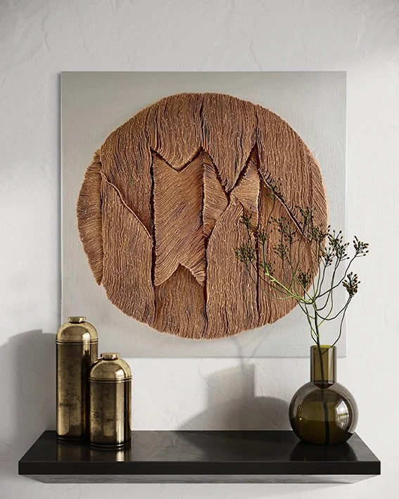 Wanddecoratie met een groot kunstwerk van een fijn gekeerde houtcirkel met krakeleffect, naast een zwarte tafel met twee gouden metallic flessen en een bruin glazen vaas met takken.