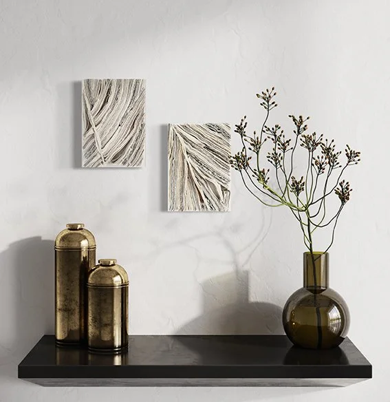 Decoratieve accessoires op een platte zwarte plank, waaronder een grote en een kleine gouden vaas, een grote groene glazen vaas met gedroogde bloemen, en twee abstracte kunstwerken aan de witte muur.