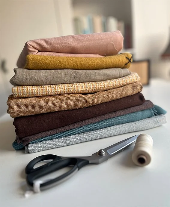 Een stapel van verschillende gekleurde stoffen, een schaar en een naald met draad op een witte tafel.