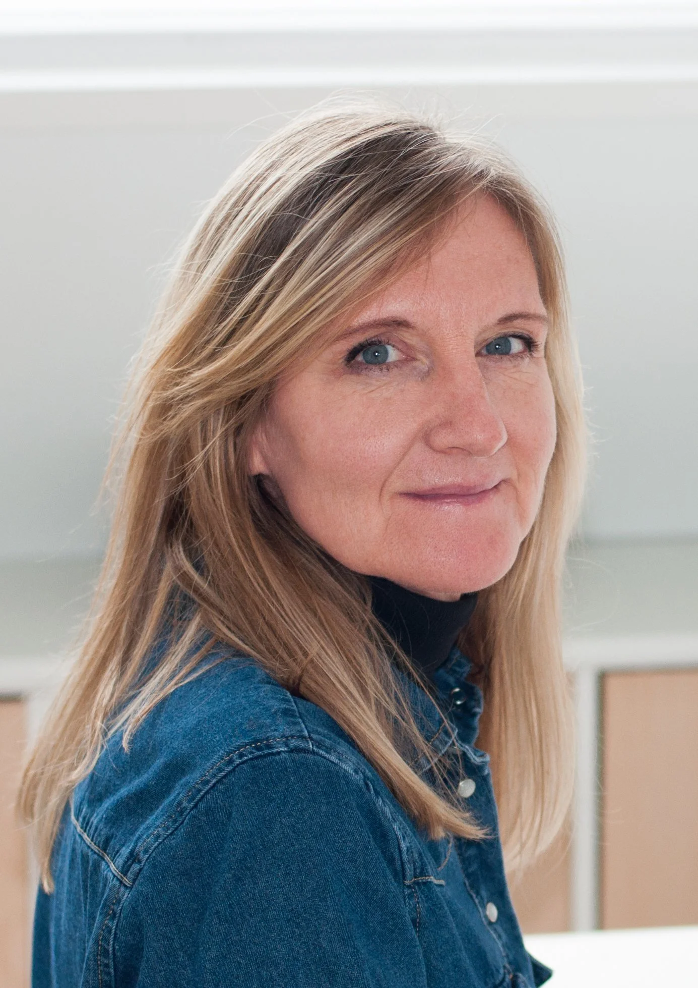 Een vrouw met blond haar, blauwe ogen, draagt een blauwe spijkerjack, zit in een kamer met witte muur en houten meubels.