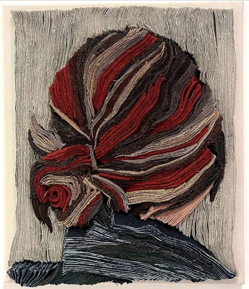 Textuurkunst van een grote bloem met rode, zwarte en beige tinten, gemaakt met geknoopte stoffen en garen.