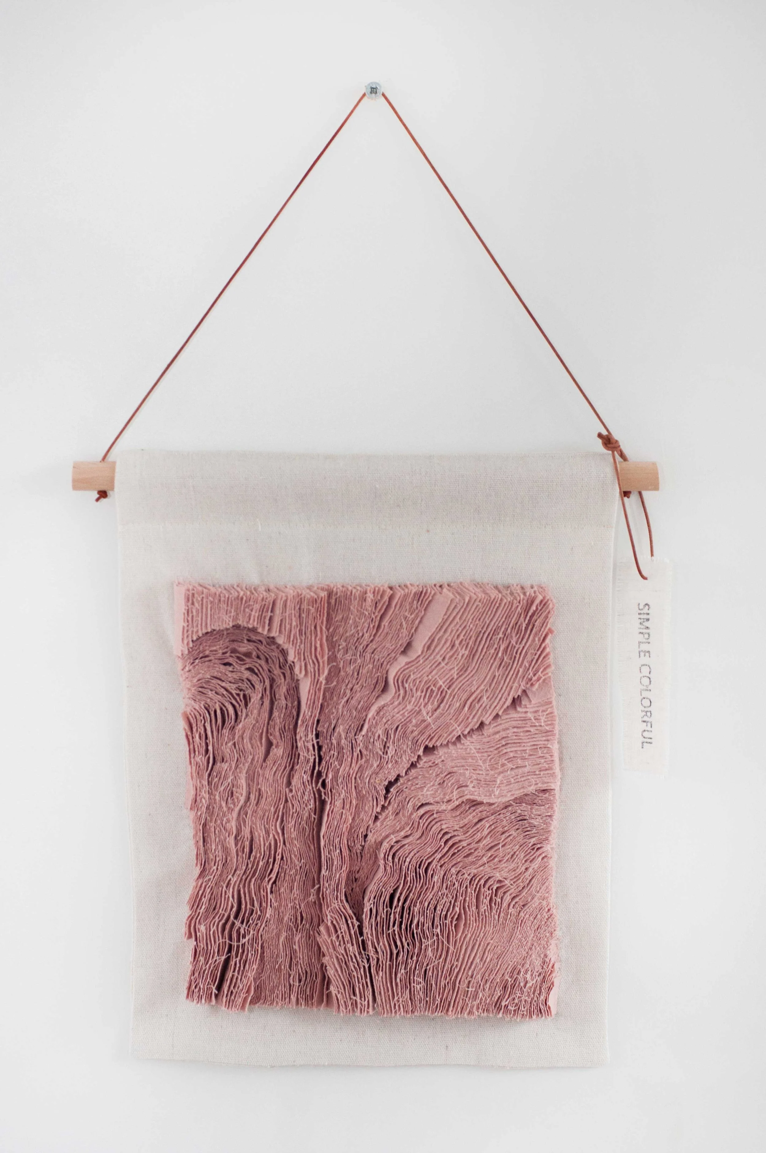 Wanddecoratie met een stoffen doek en een textuurrijke, roze geborduurde afbeelding, hangt aan een houten stok met touw aan een metalen haak tegen een witte muur.